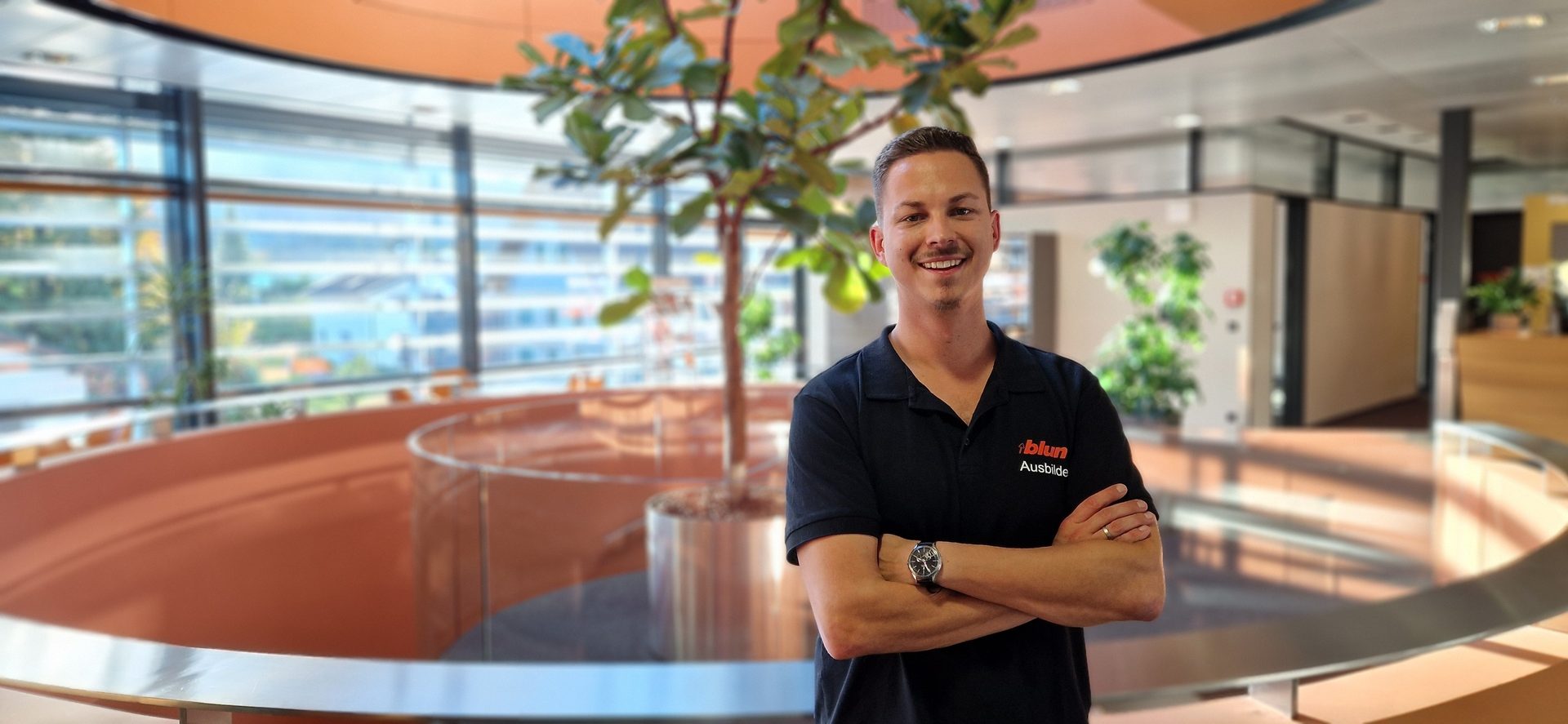 Smiling man in "blum Ausbilde" shirt, arms crossed, in a modern office with a large plant.