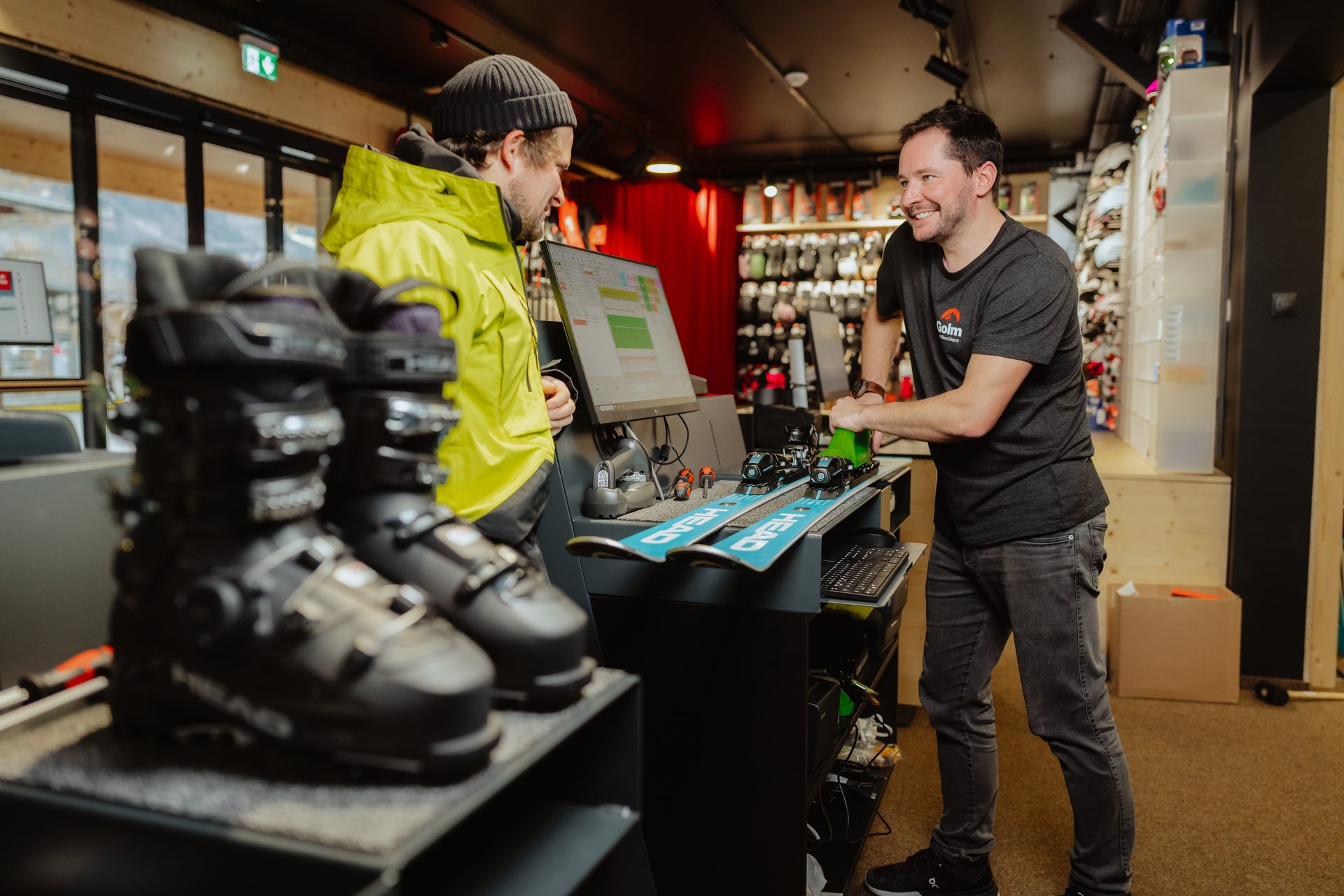 Two men in a ski shop, one customer looks at a screen, a smiling employee works on skis.