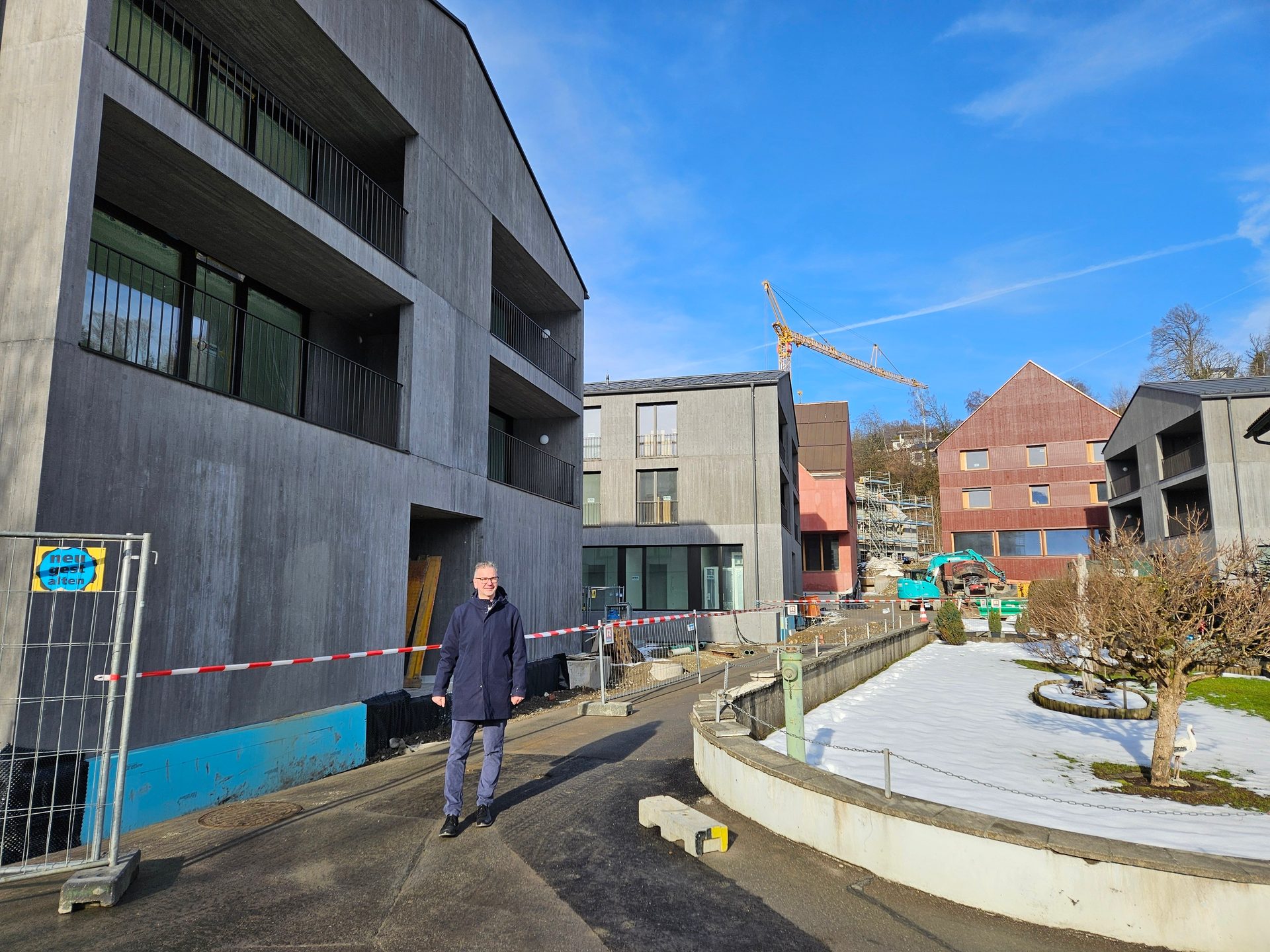 Man stands by modern concrete buildings and a construction site with snow on a sunny day.