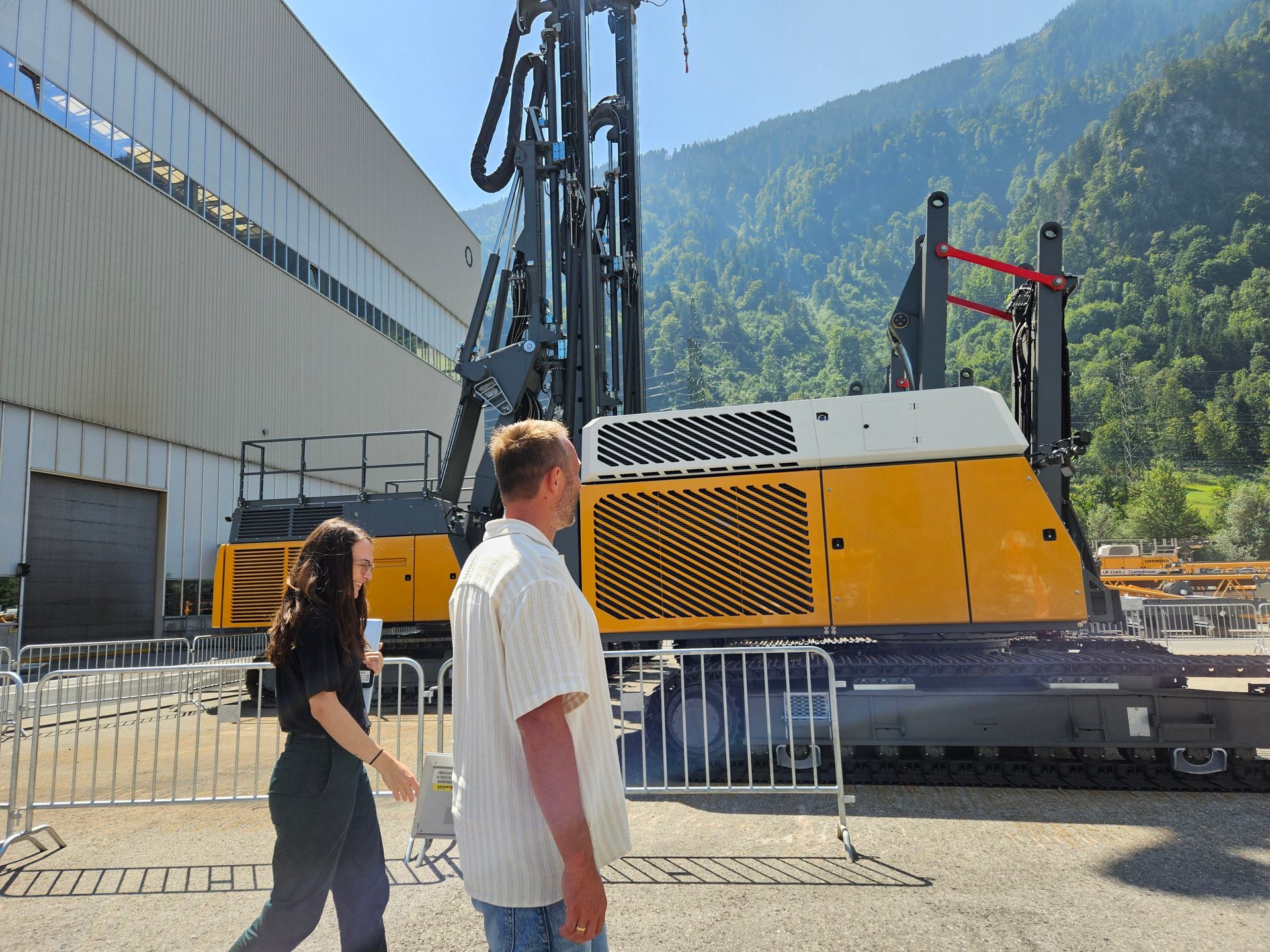 Two people walk past a large yellow drilling rig, a factory, and green mountains under a blue sky.