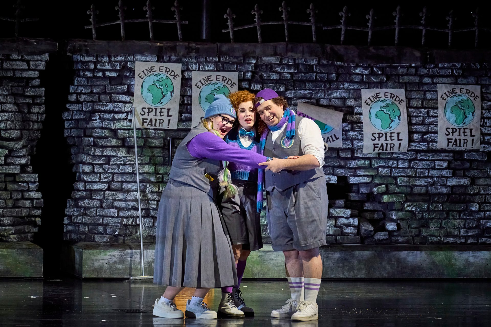 Three performers in costume, wearing purple and grey outfits, interact on a stage with brick wall backdrop and signs.