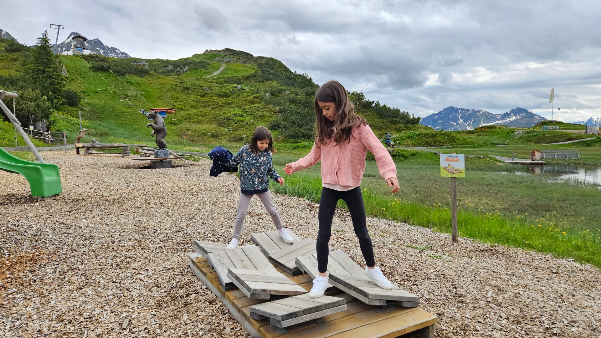 Two girls balancing on a wooden structure in a mountain playground with green hills, a lake, and a ski lift.