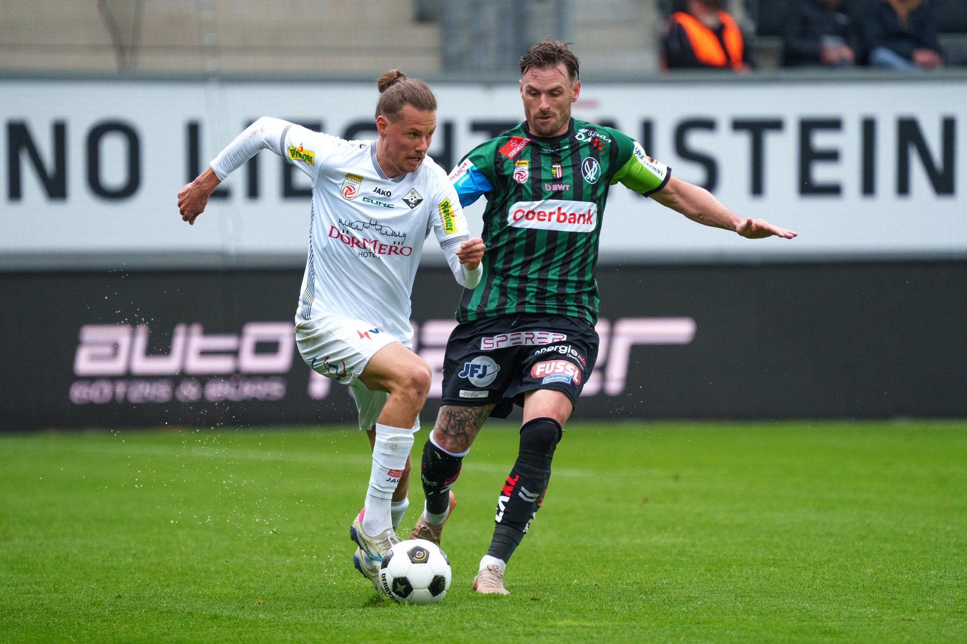 Two soccer players in white and green/black jerseys vie for the ball on a grassy field during a match.