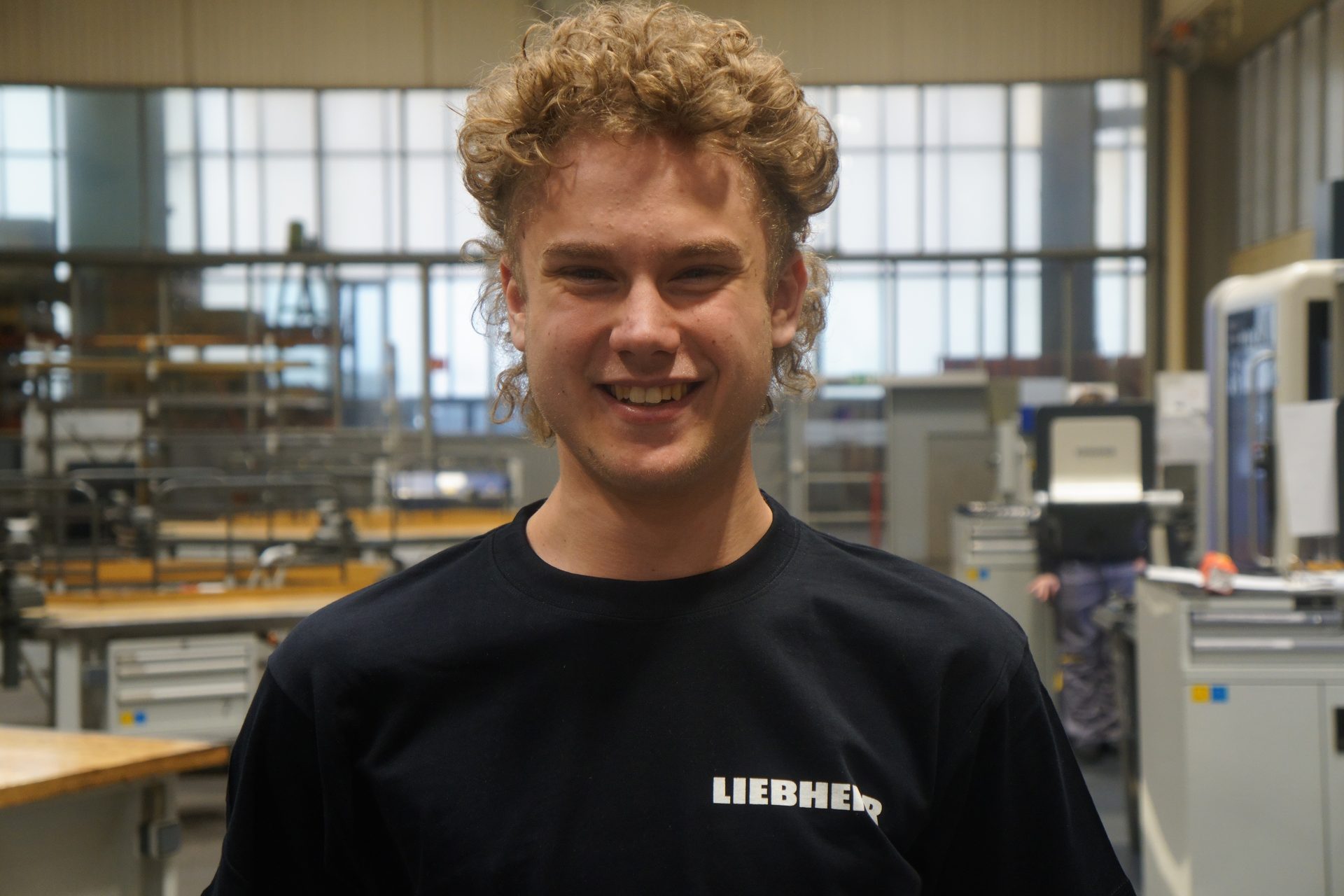 Smiling young man with curly hair wearing a Liebherr t-shirt in a workshop environment.