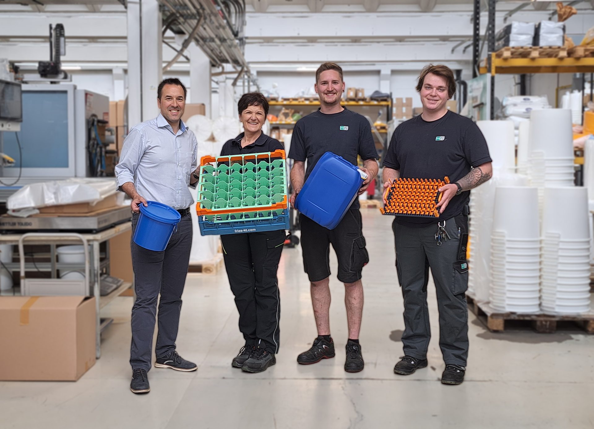 Four smiling people in a factory holding various plastic containers and crates, showcasing products.