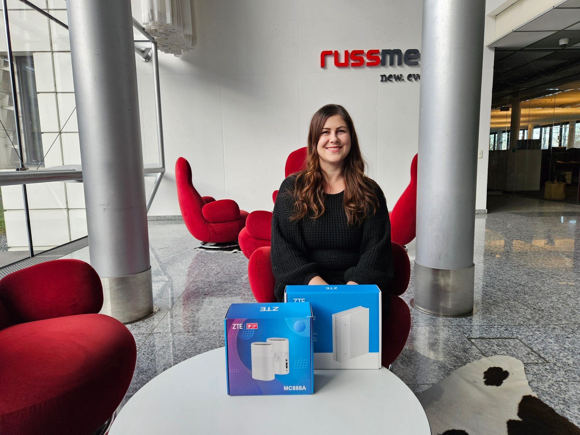 A smiling woman behind two ZTE boxes, including an MC888A, on a table in a lobby with a "russme" sign.