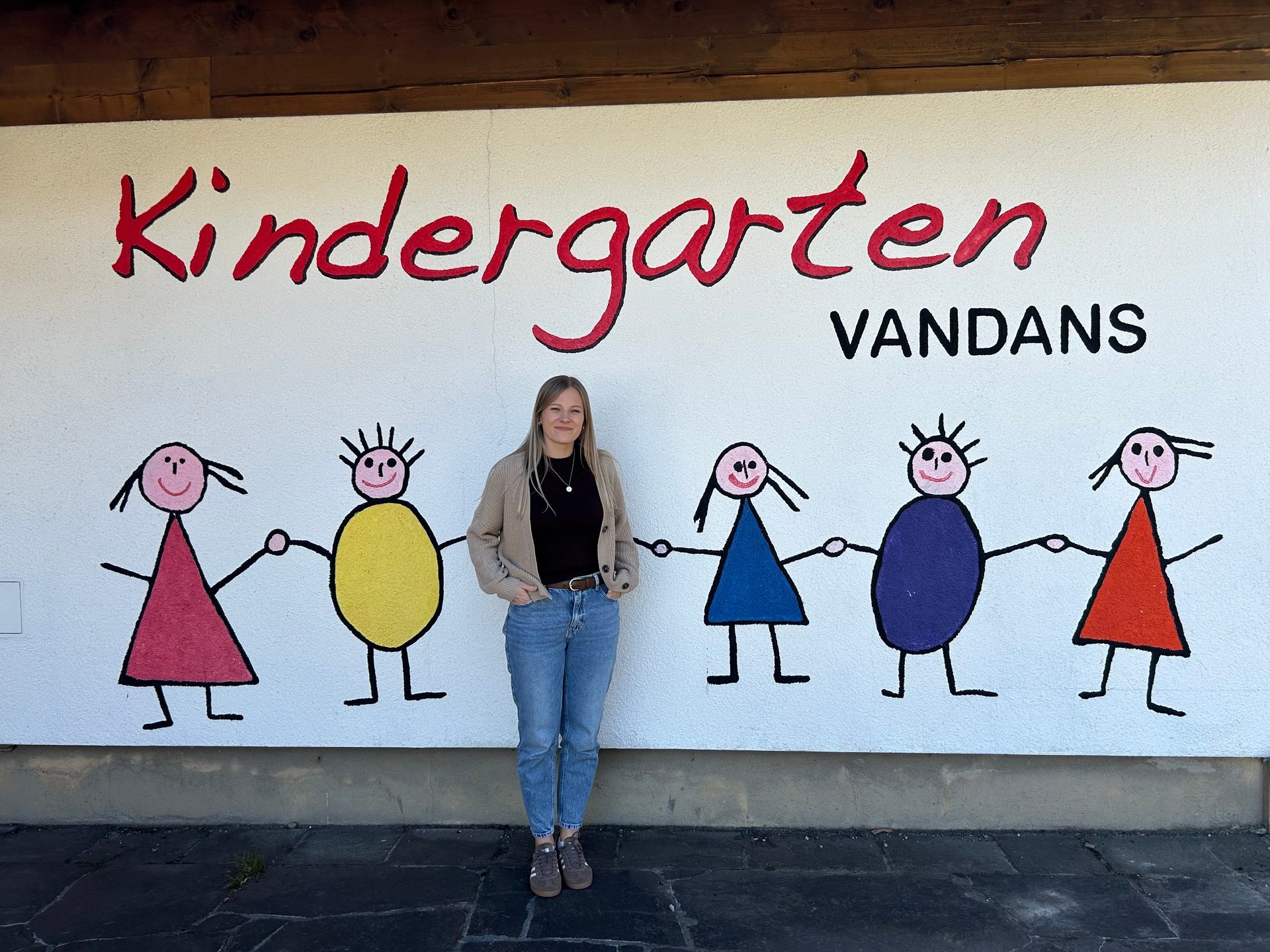 A woman stands before a wall painted with "Kindergarten VANDANS" and colorful stick figures.