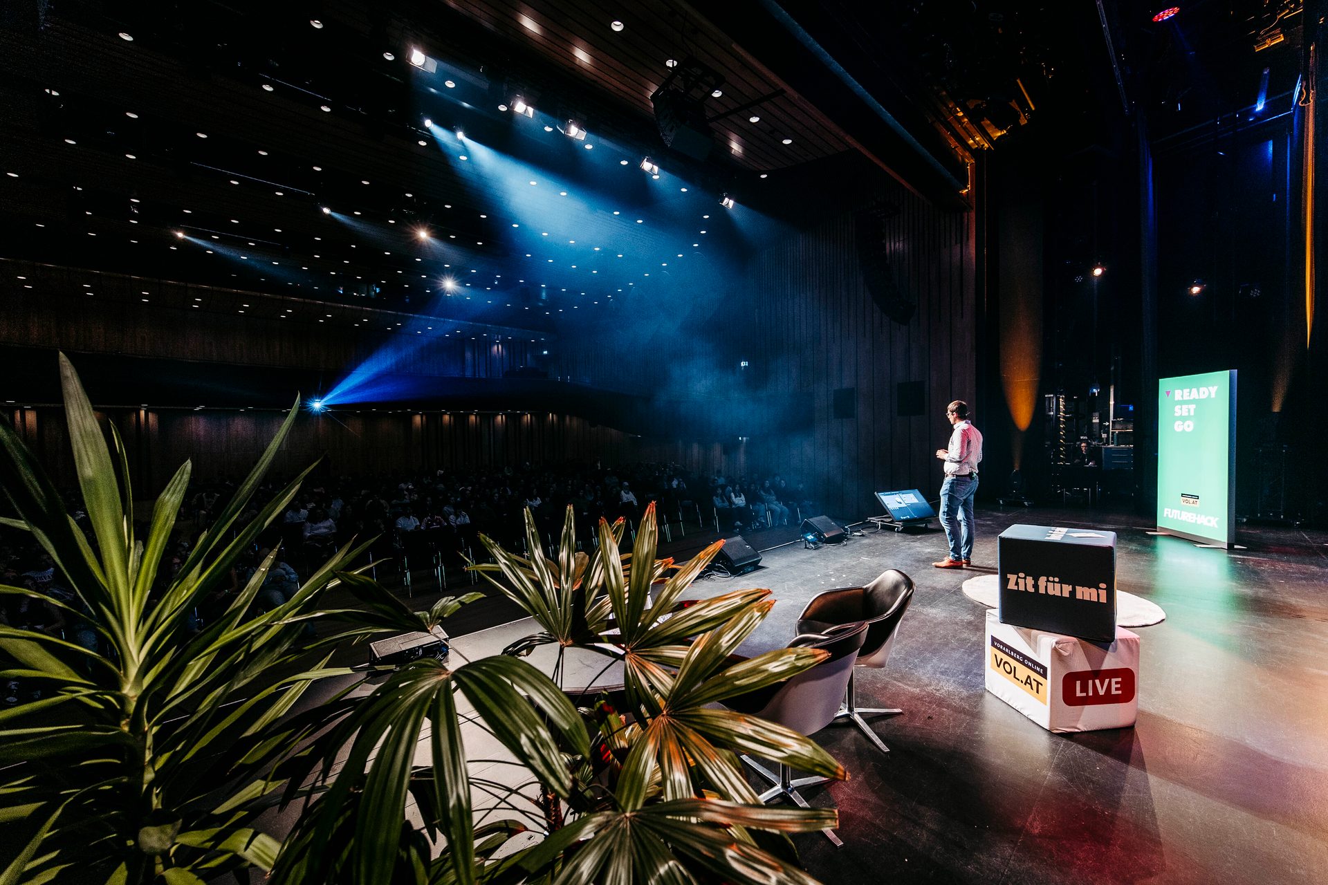 Speaker on stage with audience and spotlights, plants in foreground.
