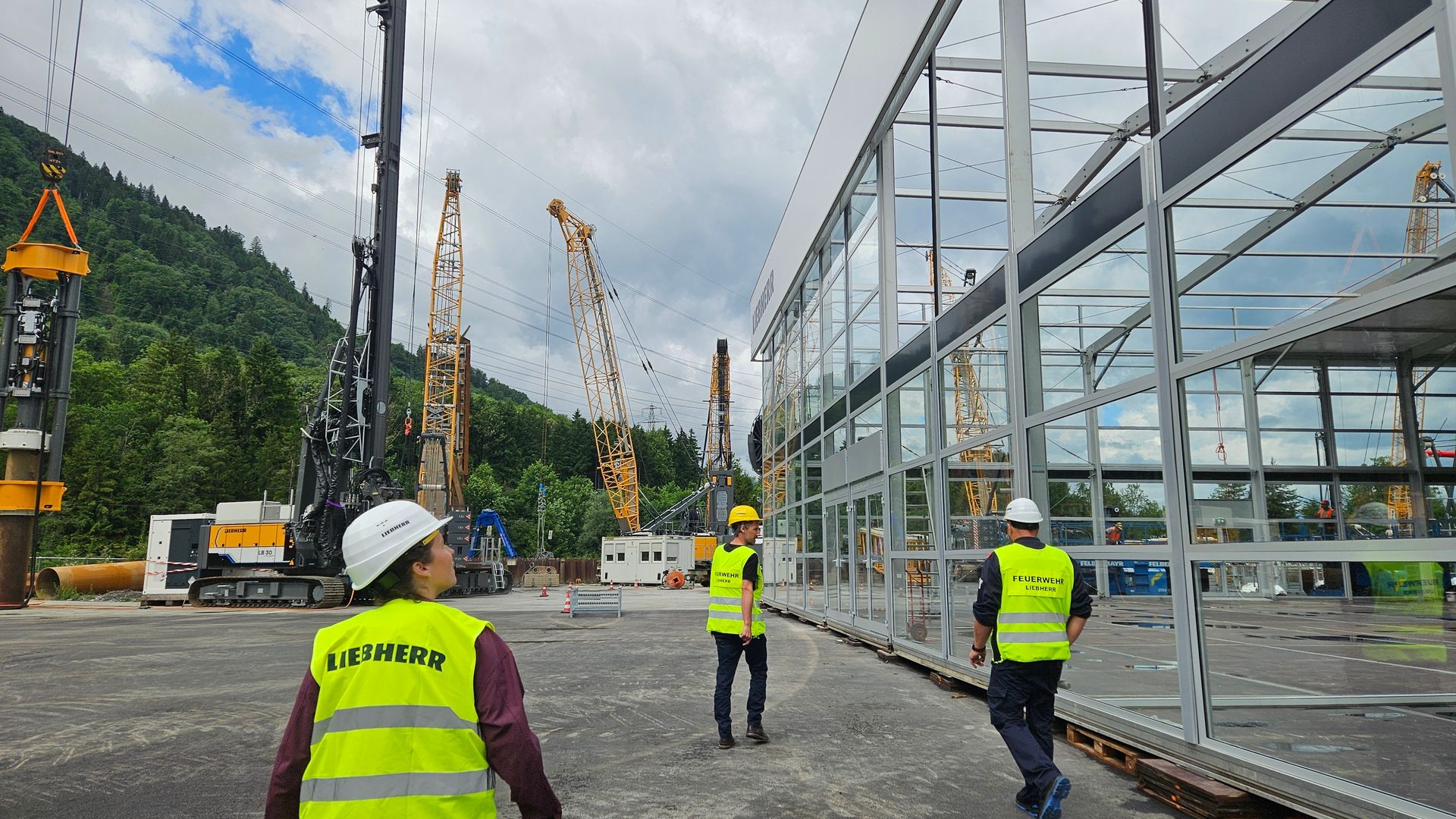 Construction site with workers in Liebherr vests, drilling rigs, and cranes, alongside a new glass building and mountains.