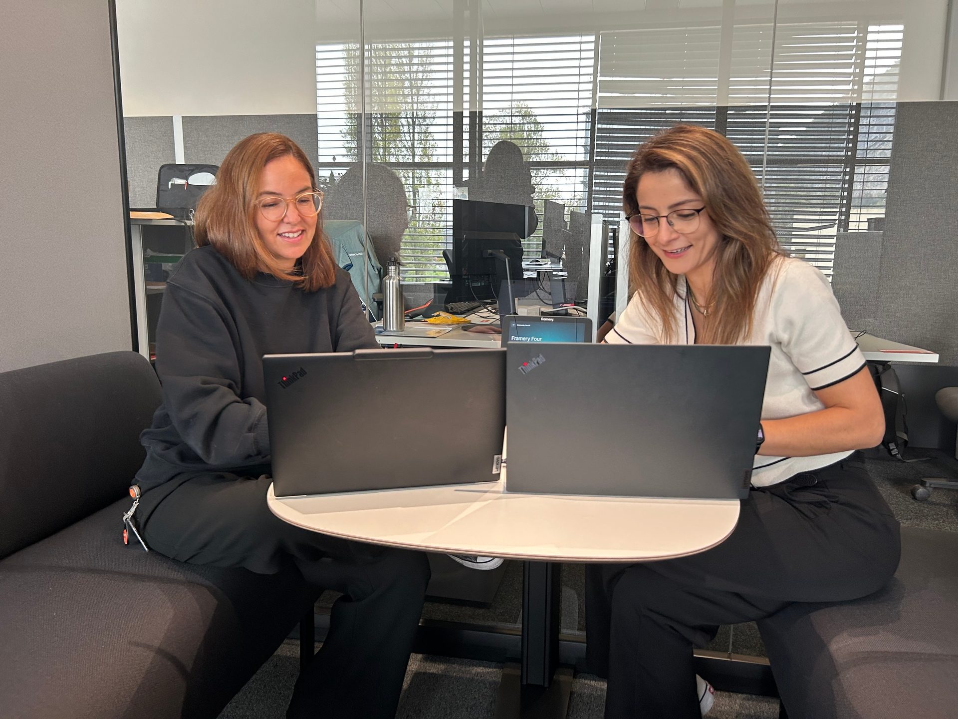 Two women smiling and working on laptops at a table in an office.