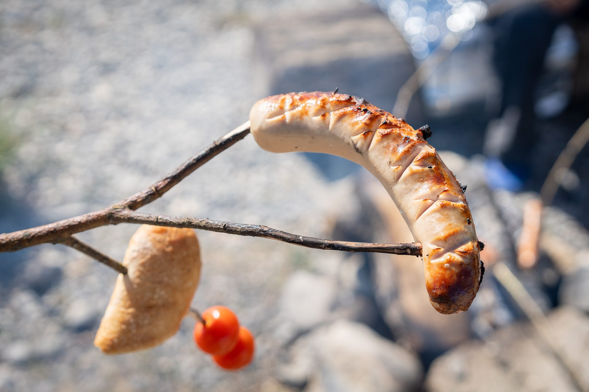 A grilled sausage, dough bread, and berries on a stick over a blurred campfire background.
