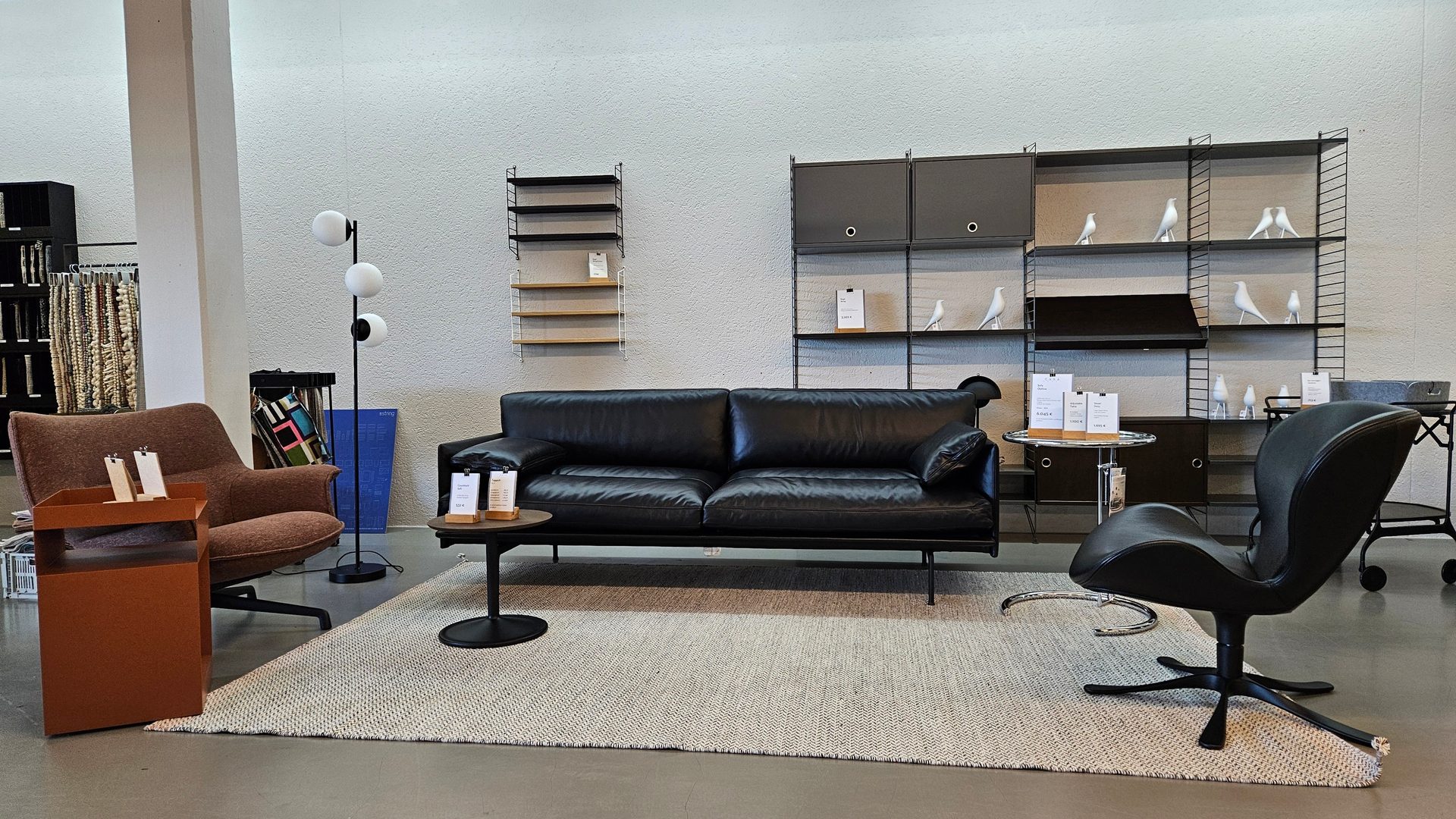 A contemporary living room setup in a store, showcasing a black sofa, armchair, swivel chair, and shelving.