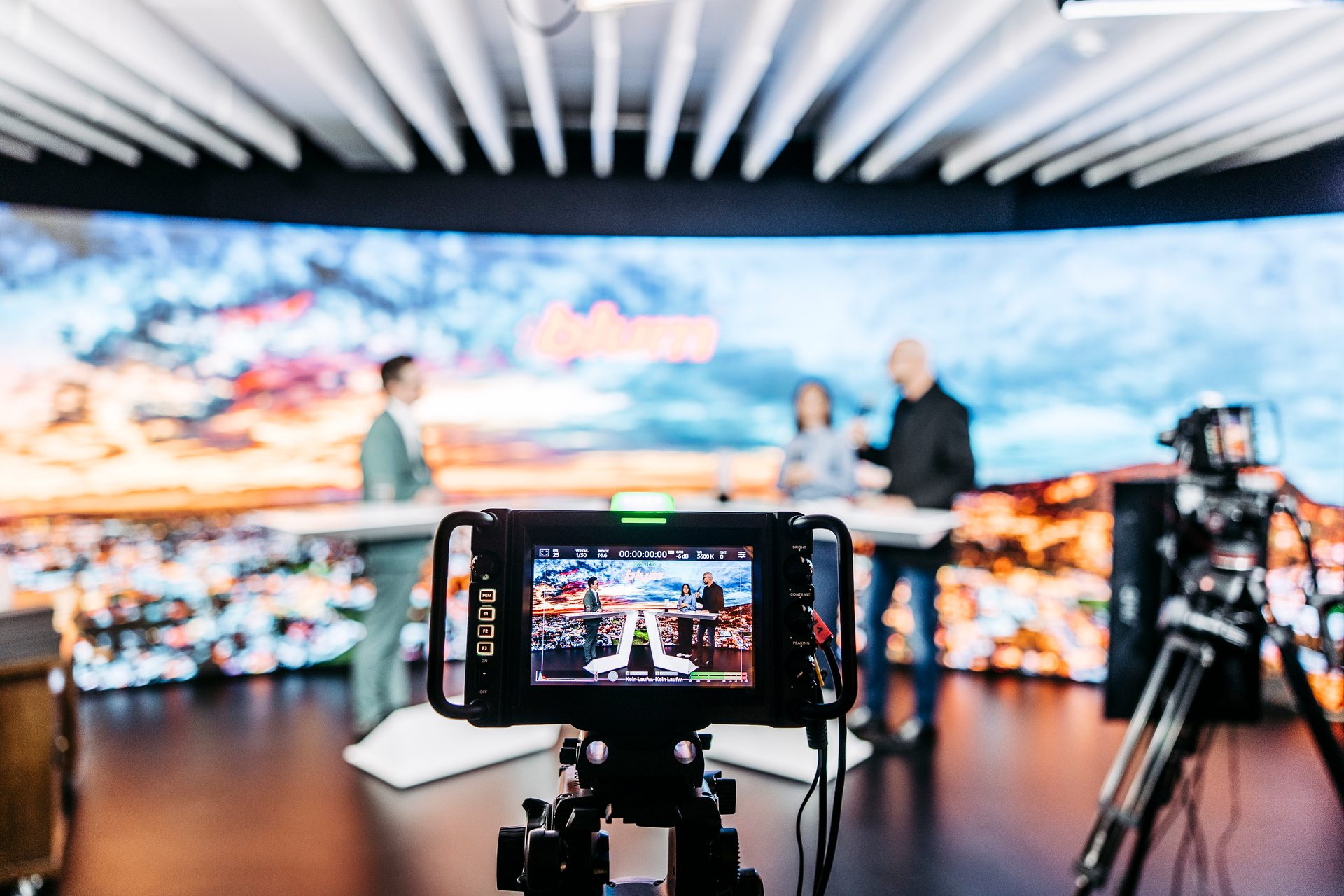 A camera monitor displaying a live broadcast of three people on a stage with an LED screen backdrop.