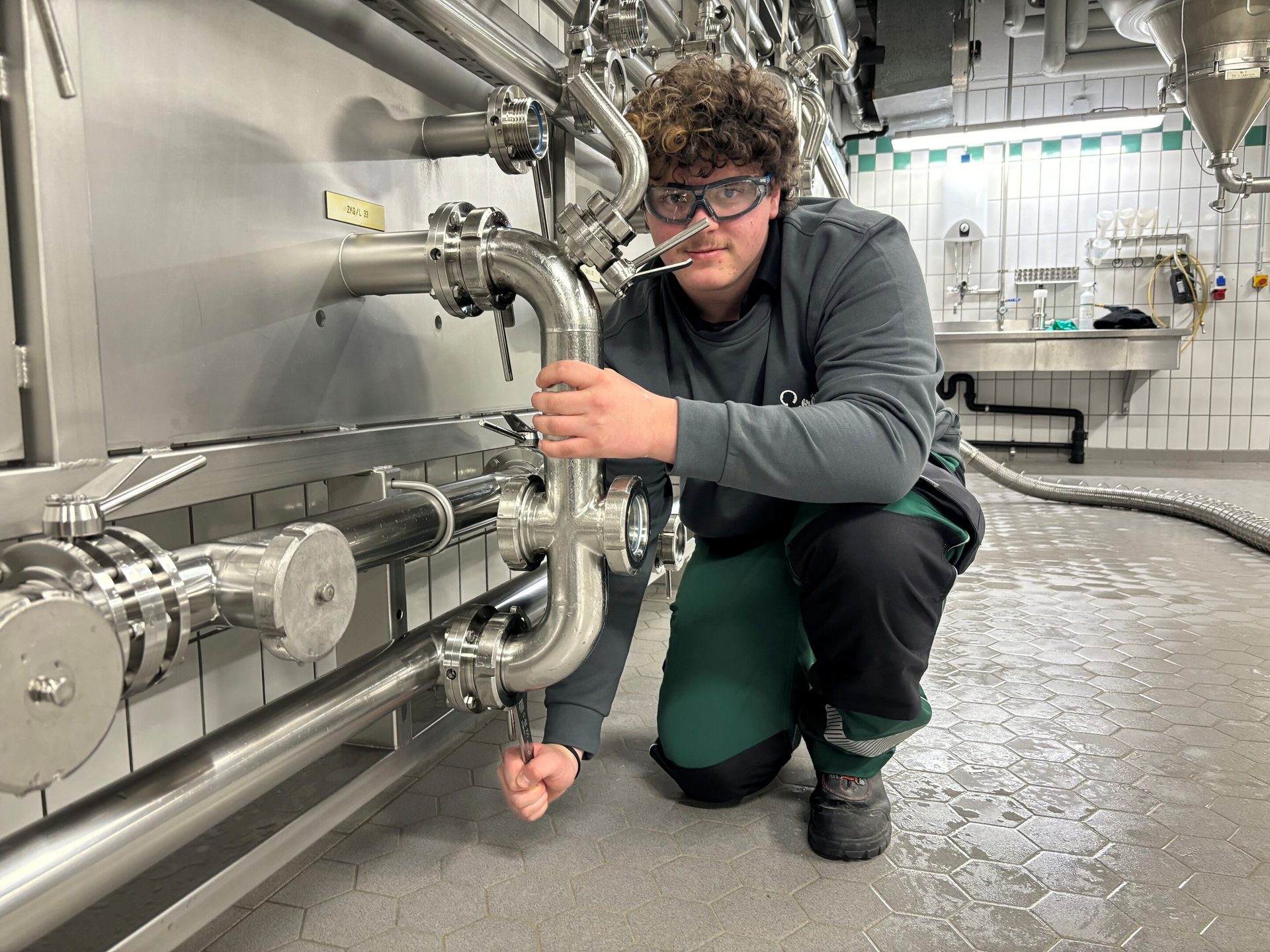 Young person in safety glasses working on stainless steel industrial pipes with a wrench.