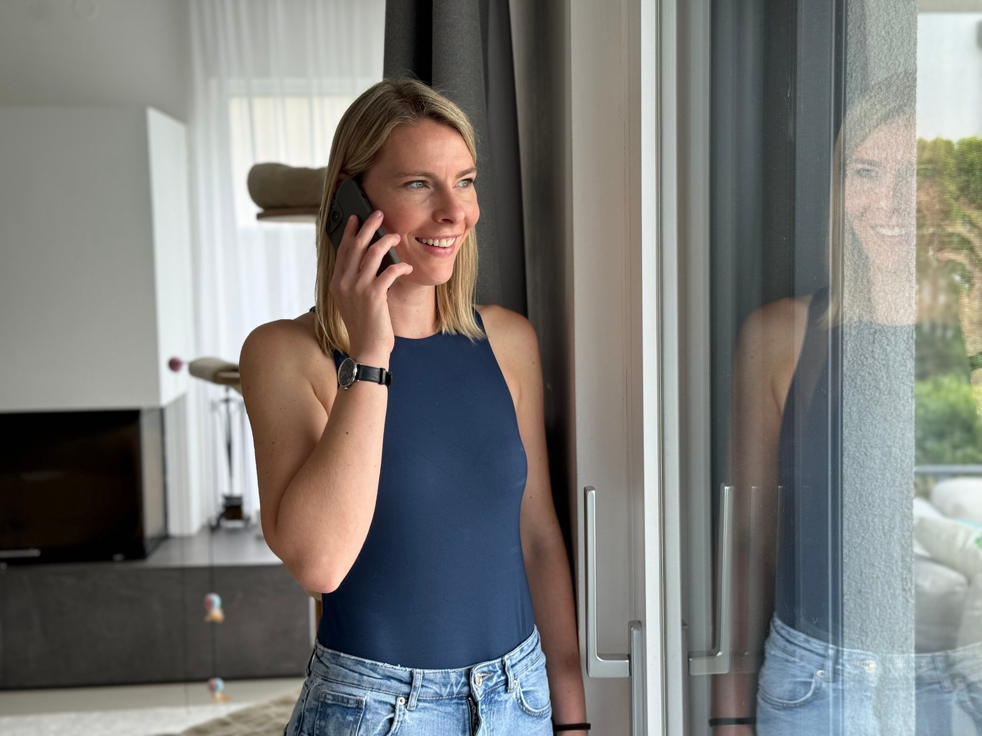 Smiling woman on phone, looking out window with reflection.