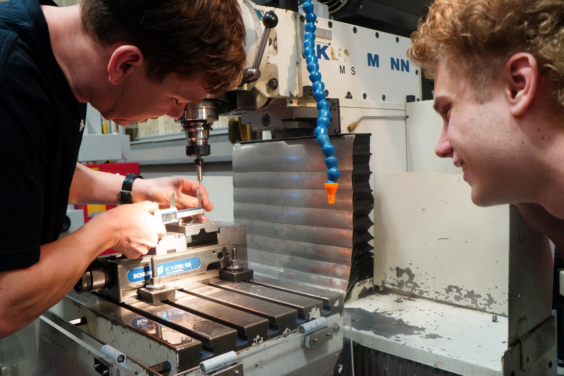Two men measuring a component on a milling machine with a caliper, one observing.