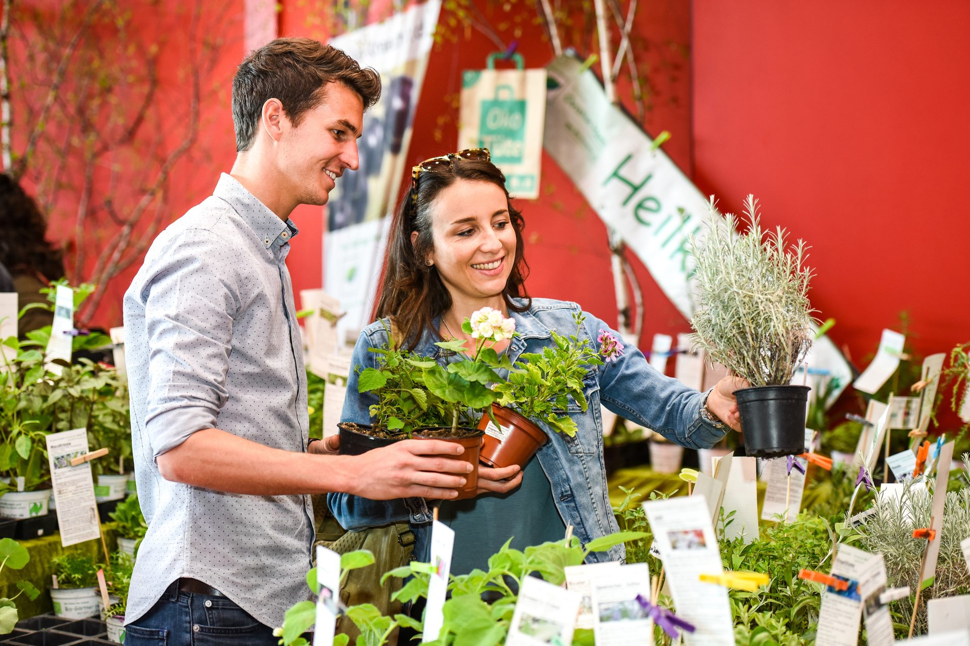 A smiling couple shops for potted plants and herbs at a market or nursery, surrounded by lush greenery.