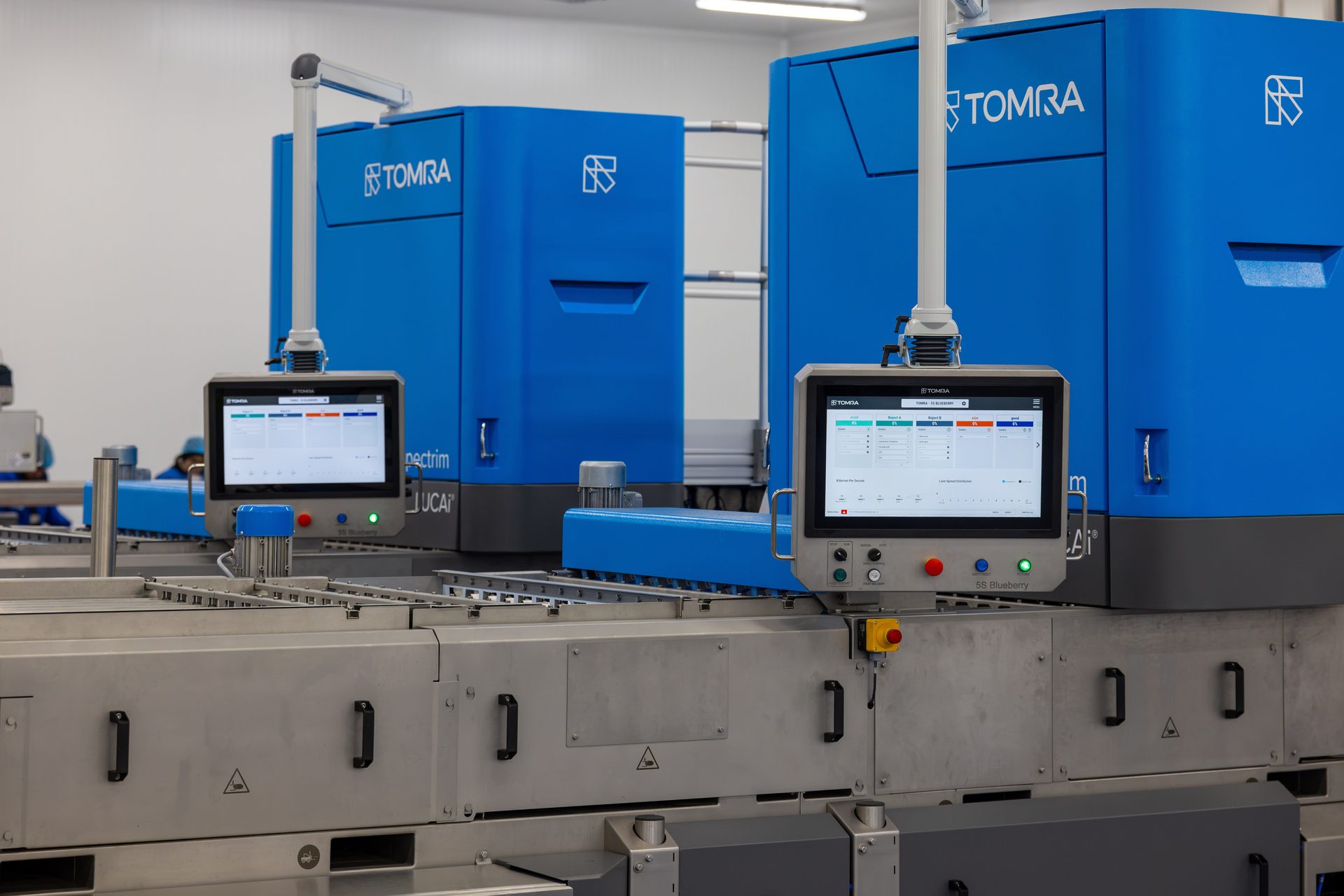 Two blue TOMRA industrial sorting machines with interactive display screens in a modern processing facility.