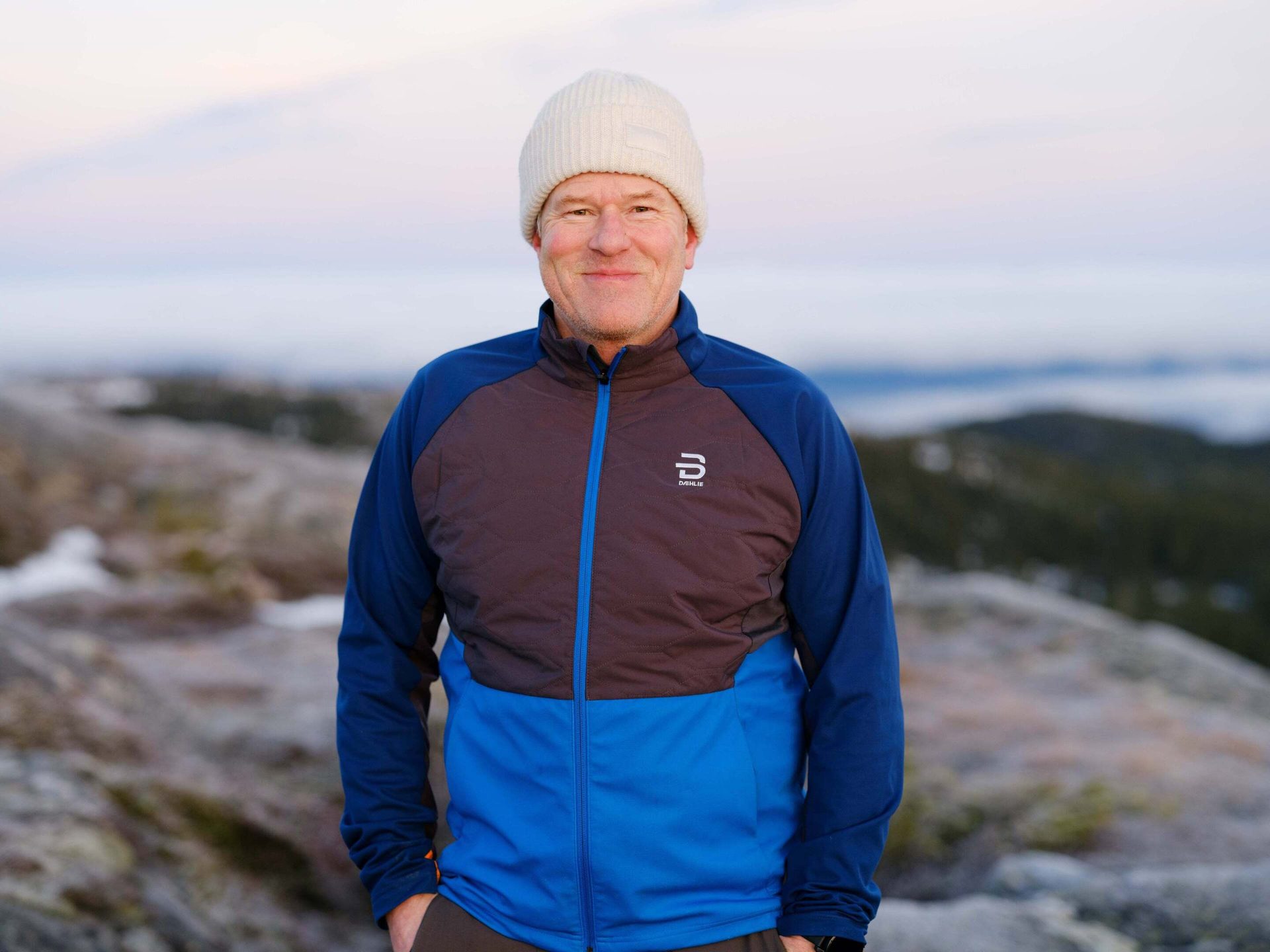 A smiling man in a blue and brown jacket and white beanie stands outdoors against a blurred landscape.