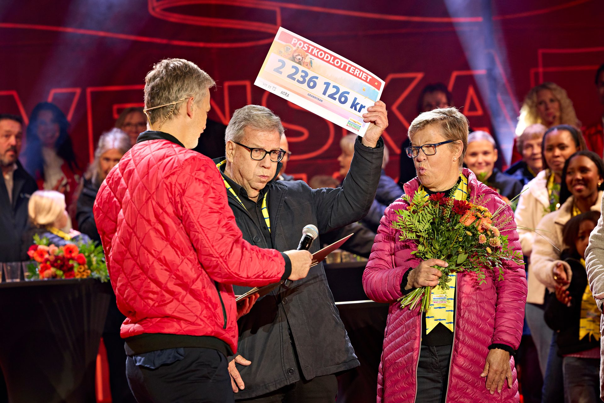 A man displays a 2.2M kr Postkodlotteriet check to a surprised woman holding flowers on stage.