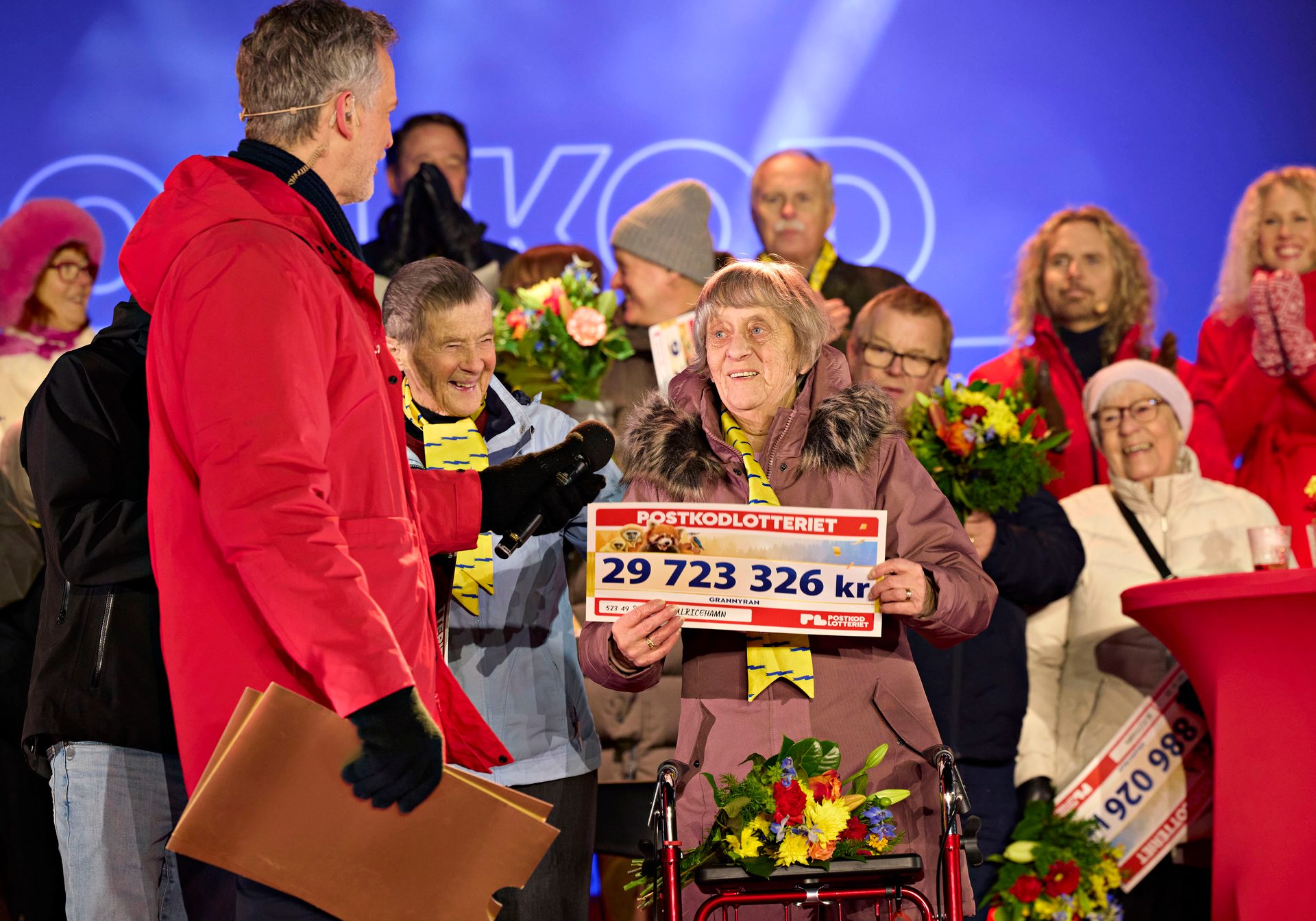 Elderly woman smiles, holding a 29.7 million SEK lottery check with a presenter on stage.
