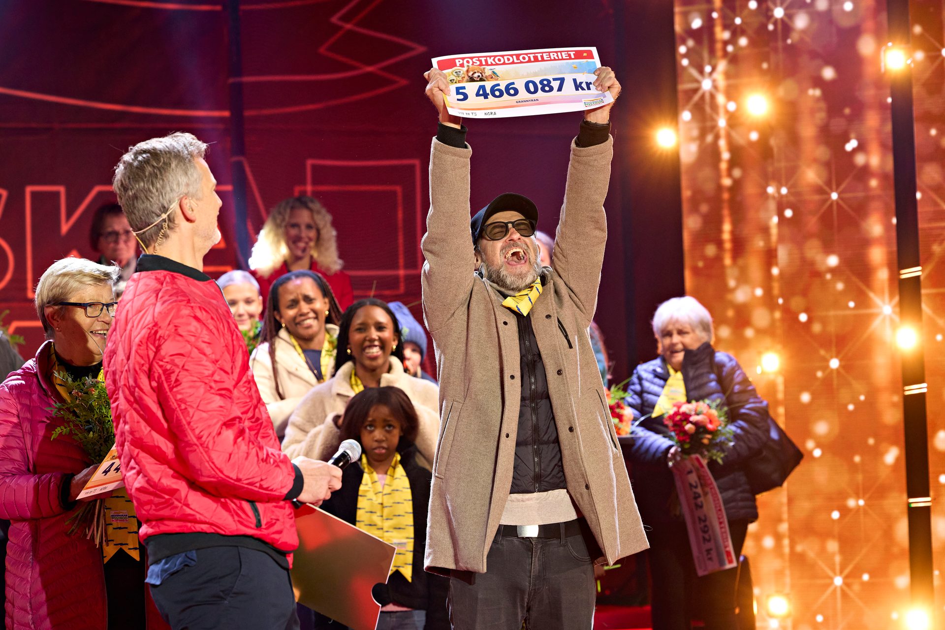 Man triumphantly holds up a giant check for 5,466,087 kr, surrounded by smiling people on stage.