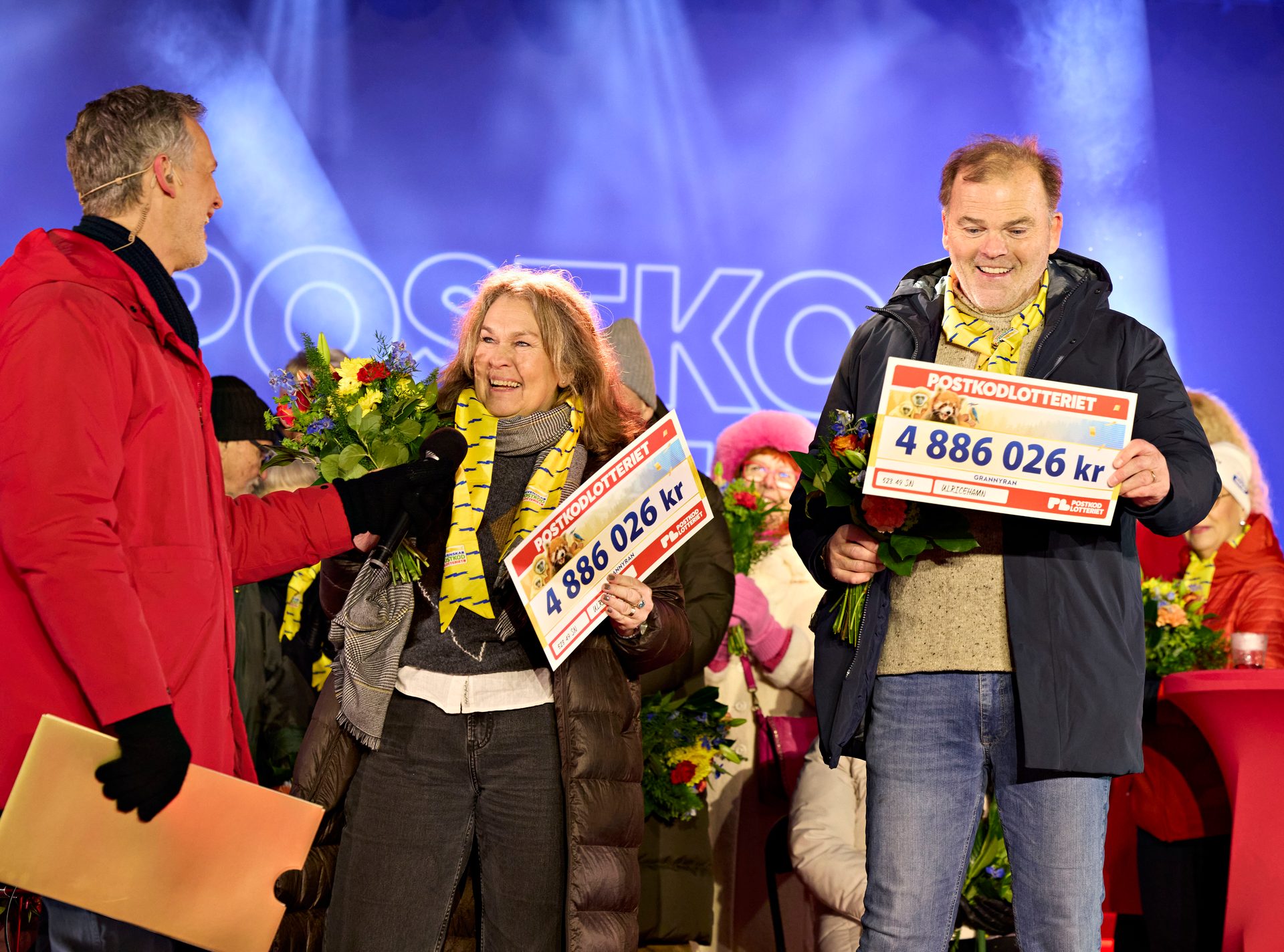 Lottery winners on stage holding large checks for 4.8 million kr and flowers, celebrating.