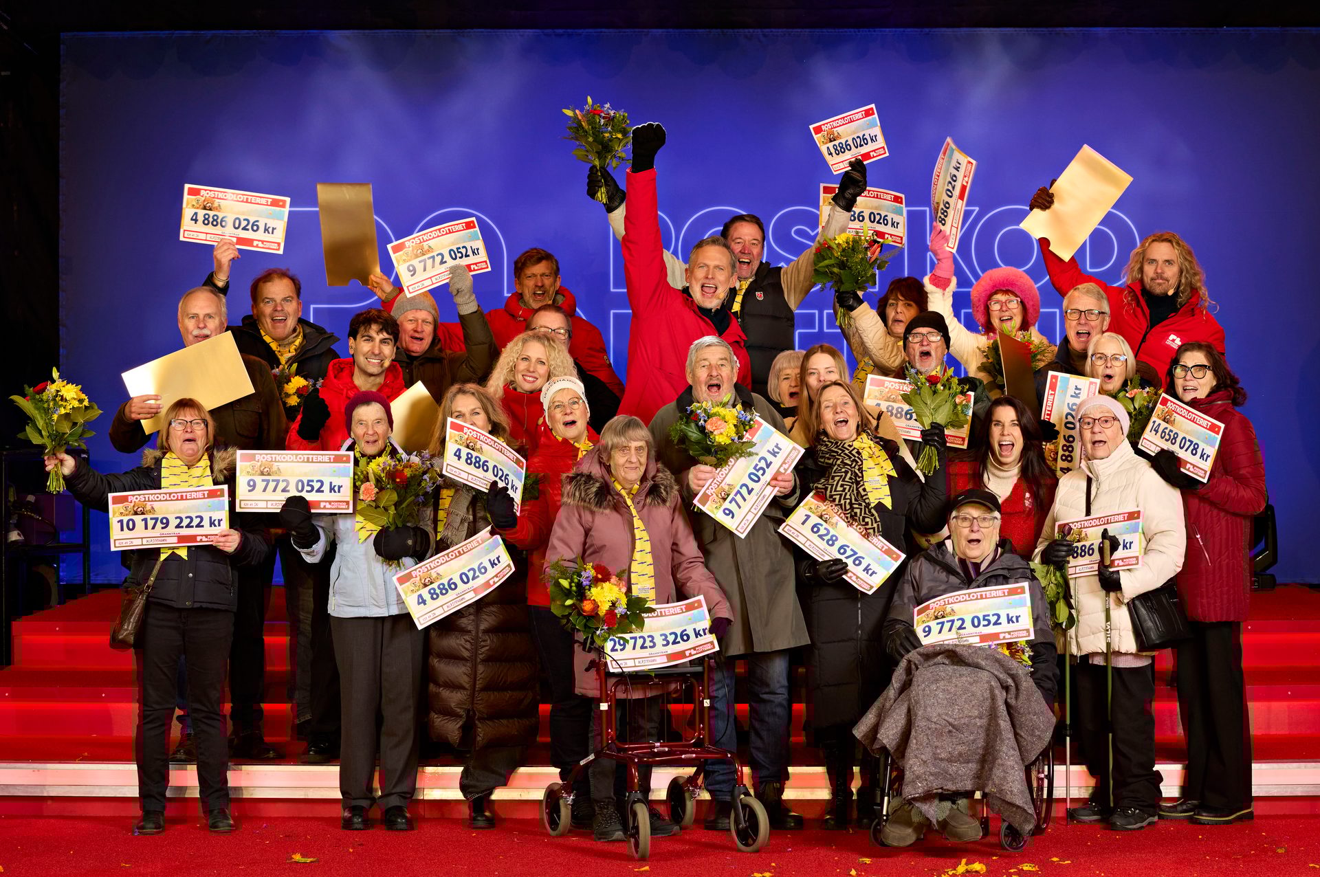 Jubilant Postkodlotteriet winners, mostly older adults, holding large checks on a stage.