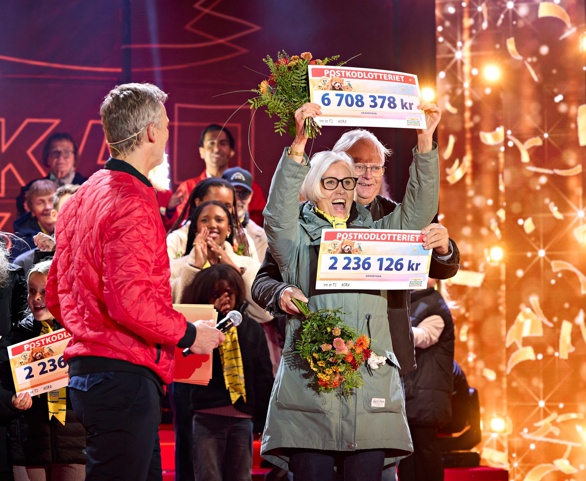 Excited lottery winners on stage hold large prize checks and flowers, with a host nearby.