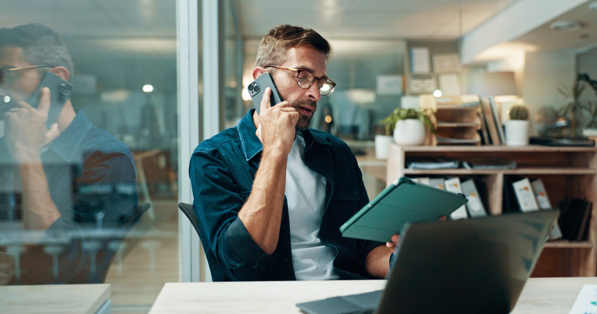 Man on phone, looking at tablet, with laptop on desk in an office.