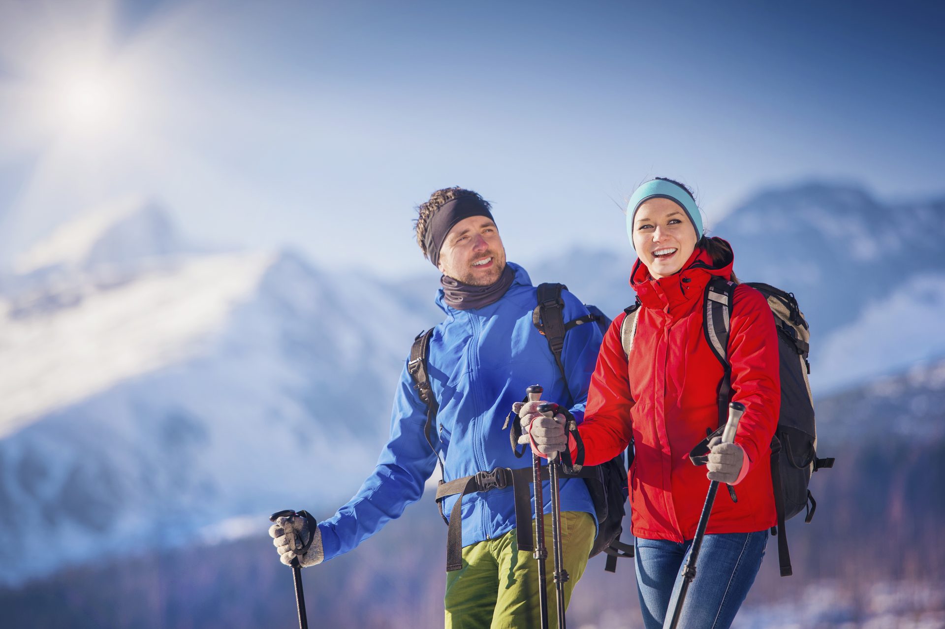 Two smiling winter hikers with poles and backpacks in sunny, snow-covered mountains.