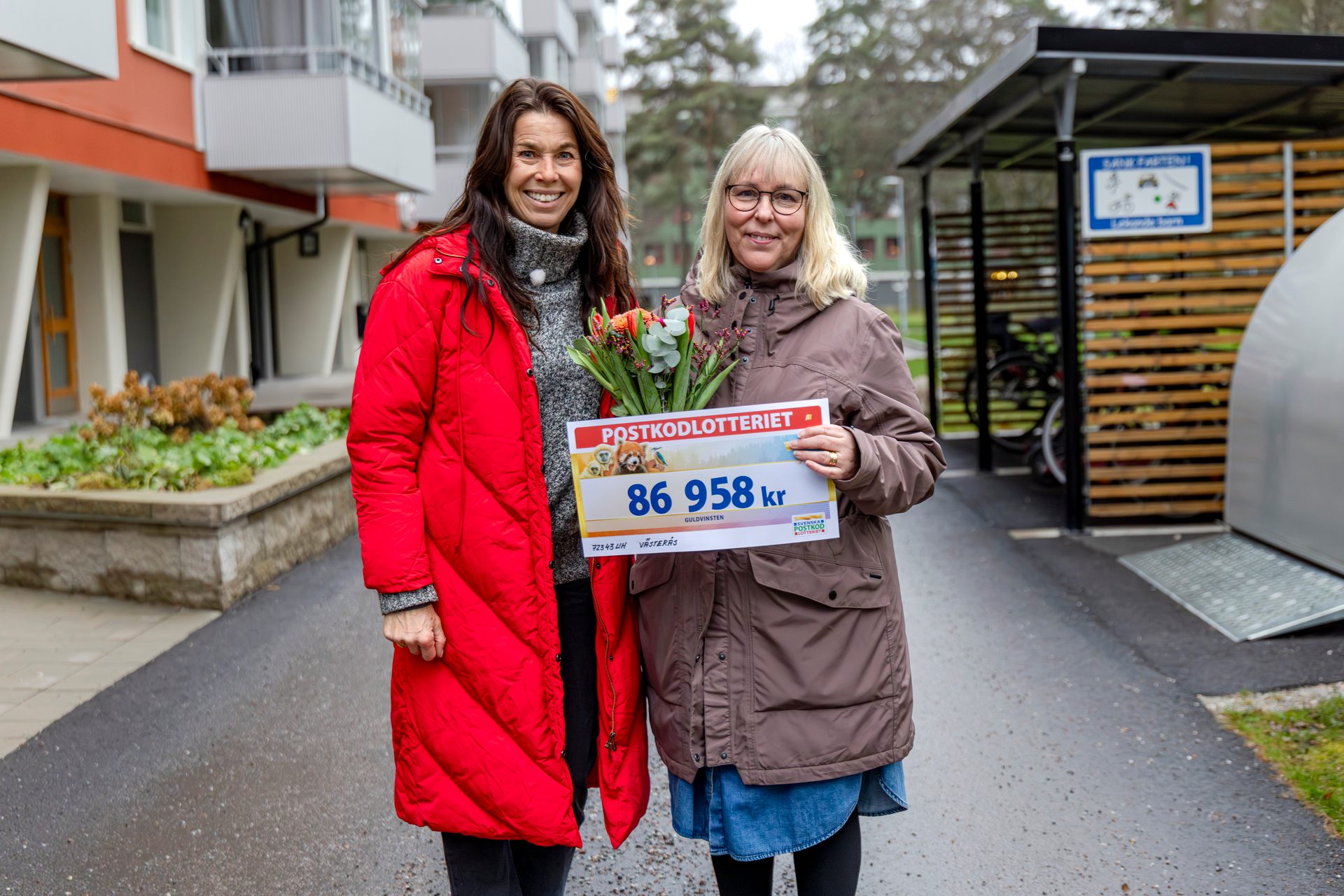 Two smiling women hold flowers and a large check for 86,958 kr from PostkodLotteriet outdoors.