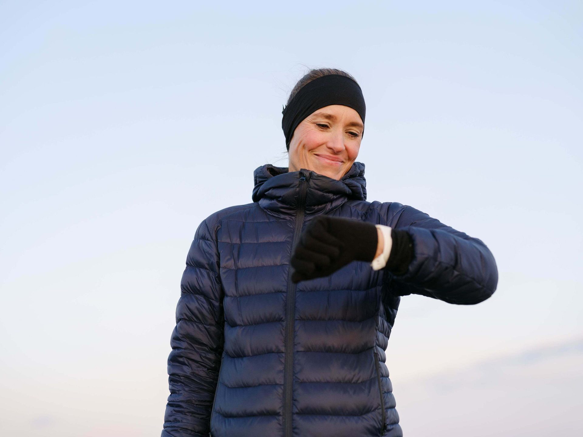 Smiling woman in a dark blue jacket and black headband looks at her wrist.