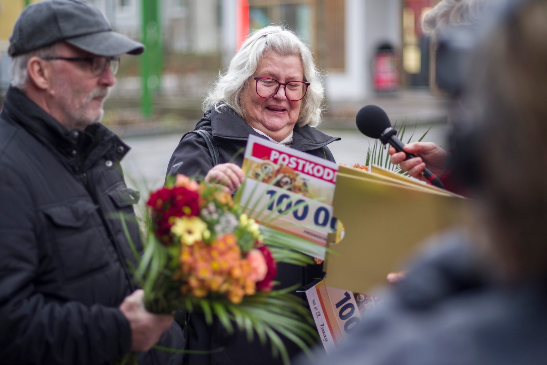 Smiling woman holding "POSTKODE 100 00" sign and flowers, next to a man, being interviewed.