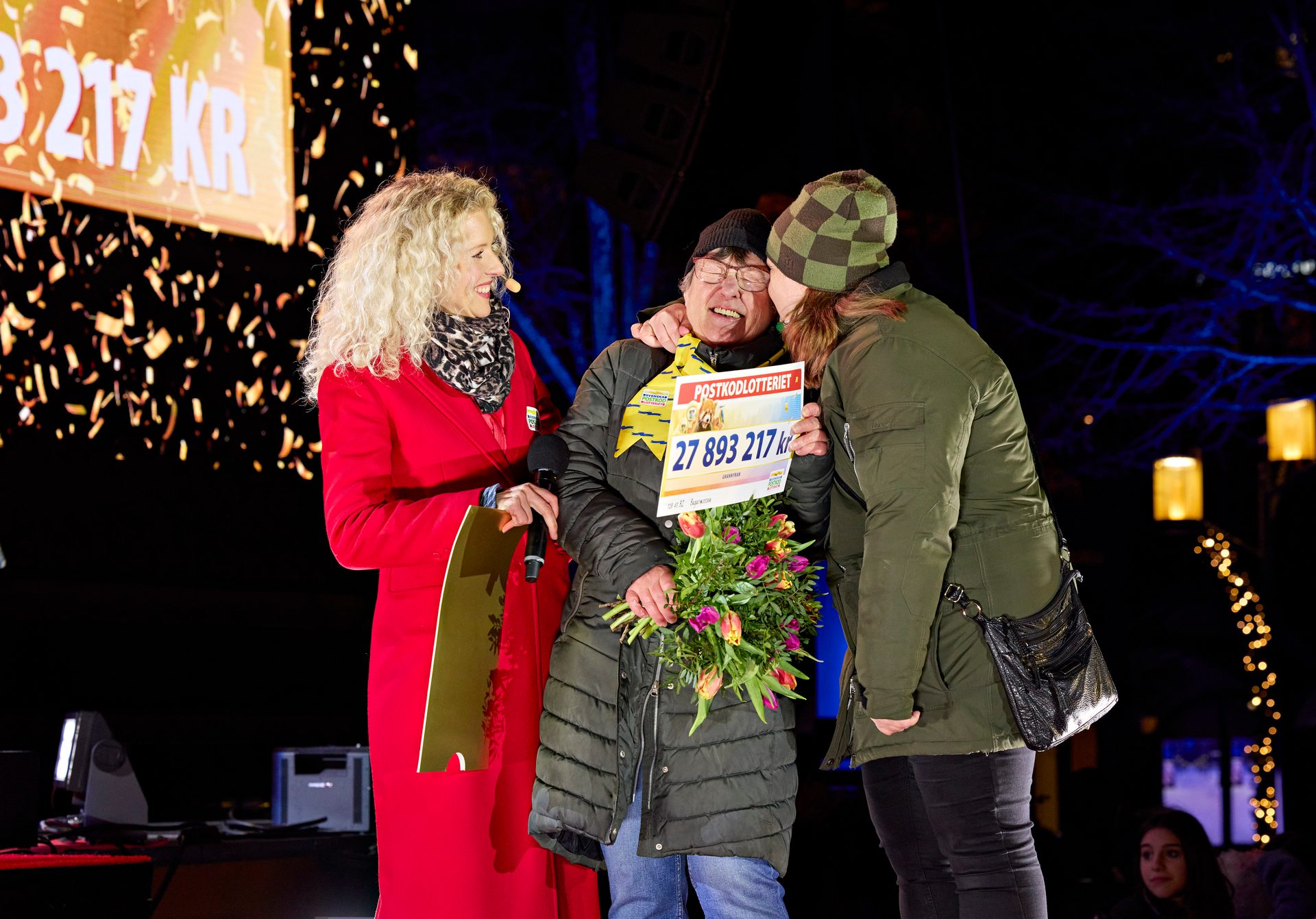 Lottery winner holds giant check for 27.8 million KR and flowers on stage, hugged, confetti falling.