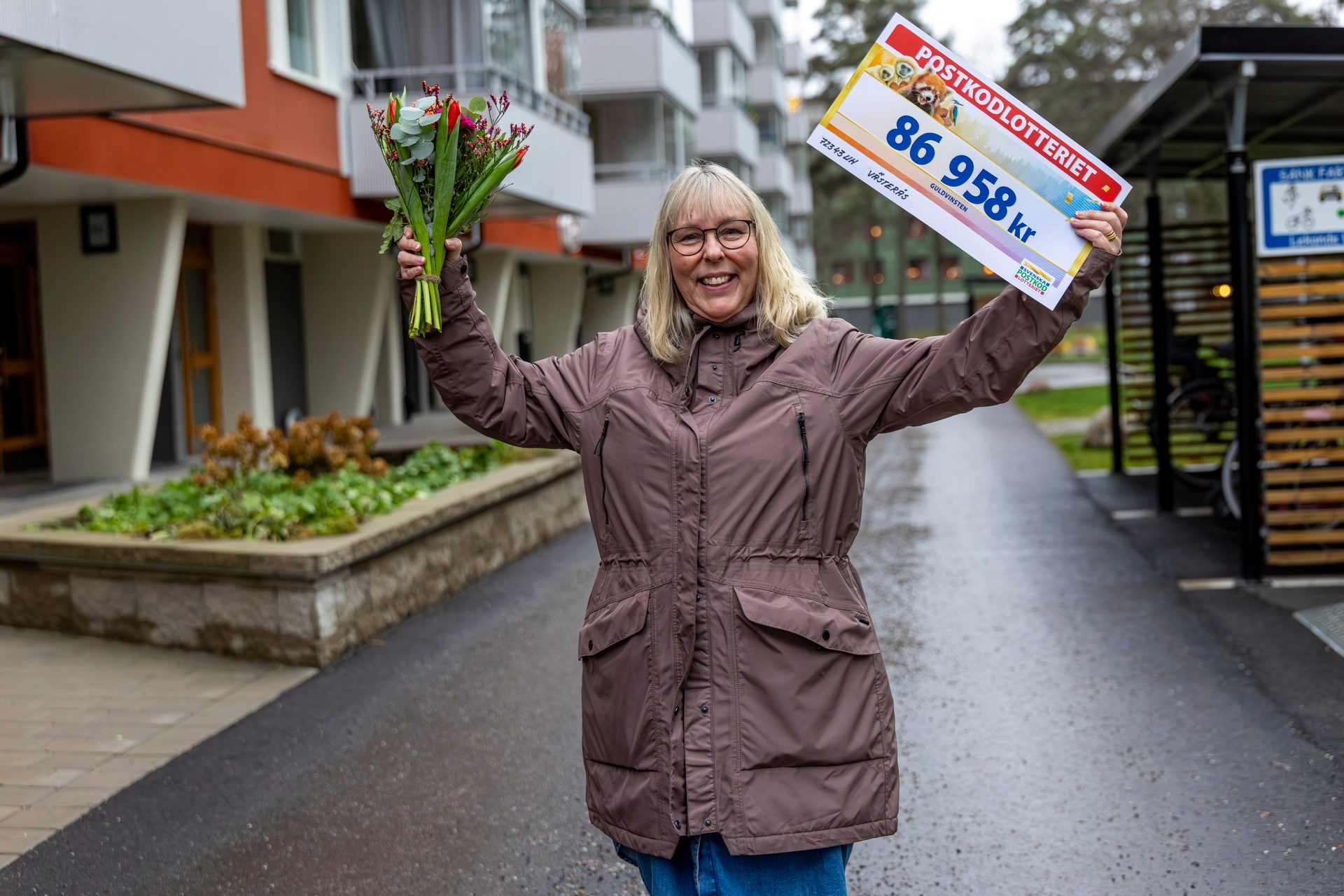 A smiling woman holding a bouquet of flowers and a large lottery check for 86,958 kr.