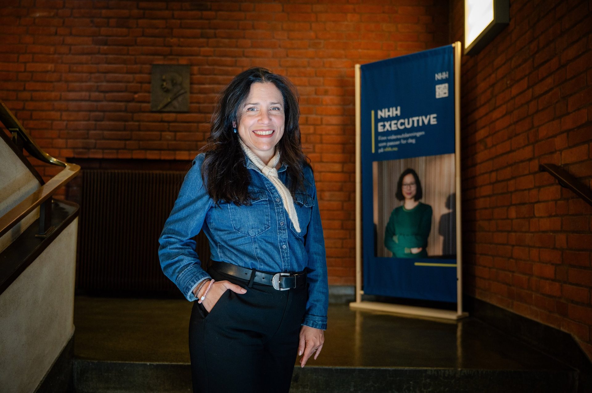 Smiling woman in denim shirt indoors, brick wall, NHH Executive sign.