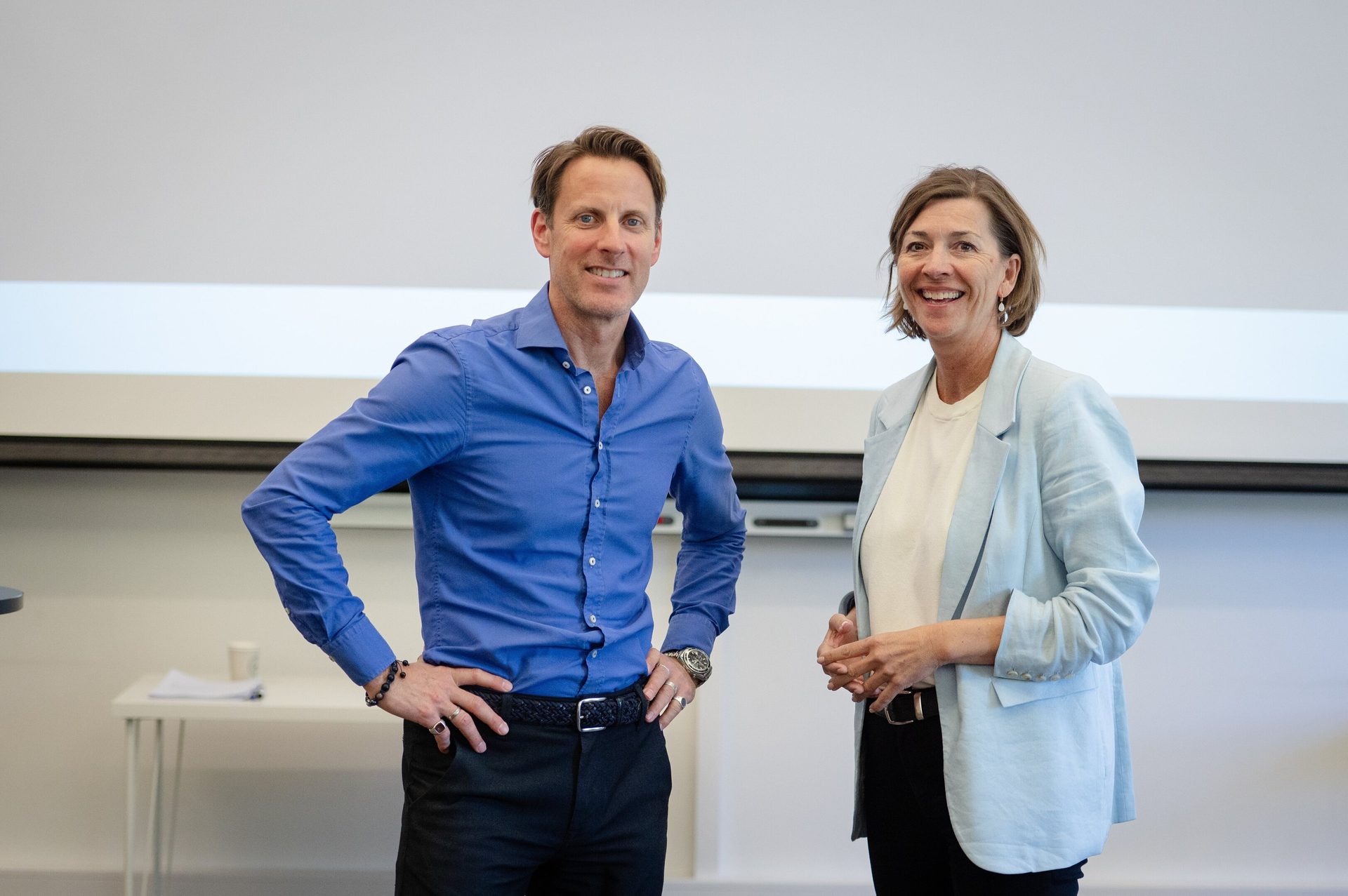 Two smiling people, a man in a blue shirt and a woman in a light blue blazer, stand in a bright room.