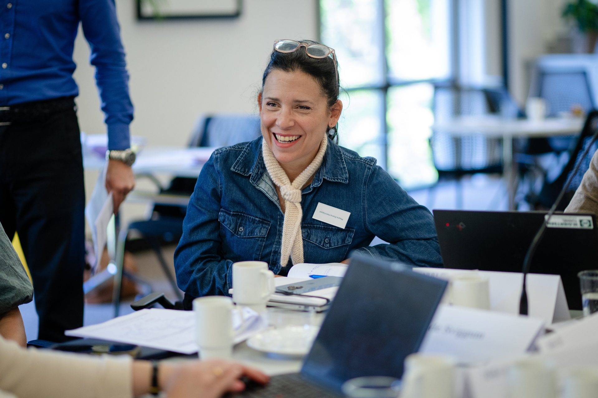 Smiling woman in denim shirt with glasses on head at a meeting table.