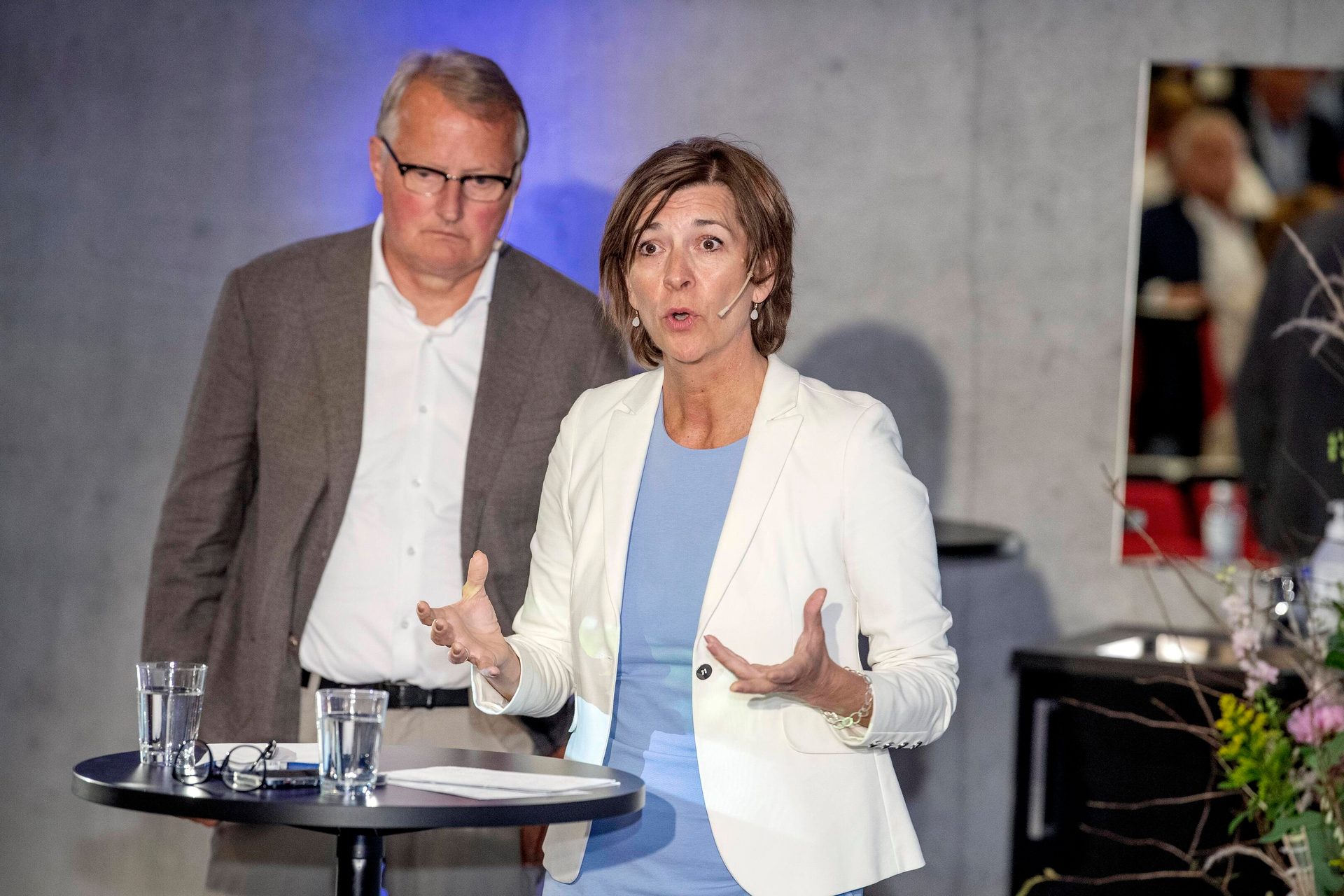 Woman speaking, gesturing, with a man standing behind her next to a table with glasses.