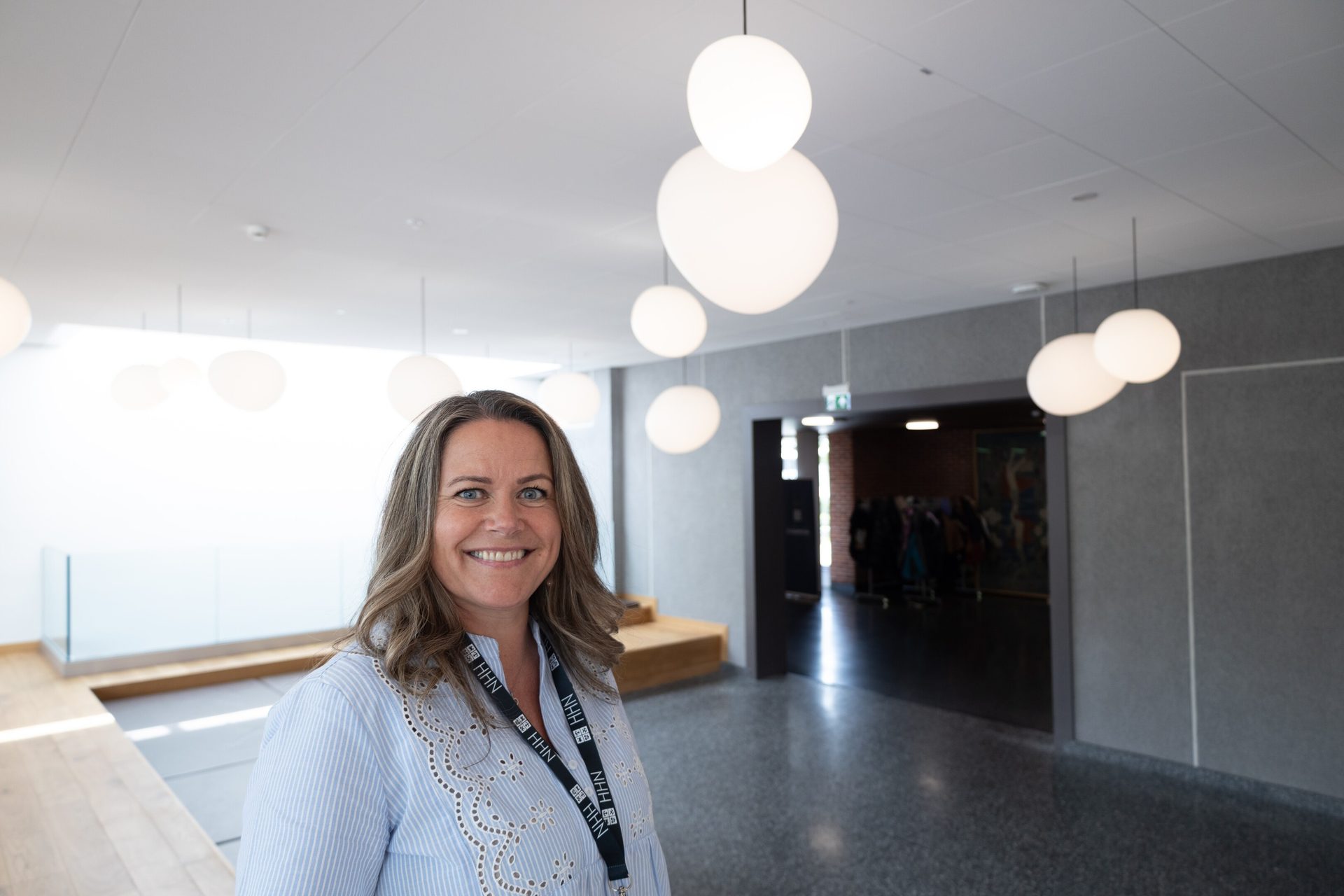 Smiling woman with NHH lanyard in a modern building with hanging lights.