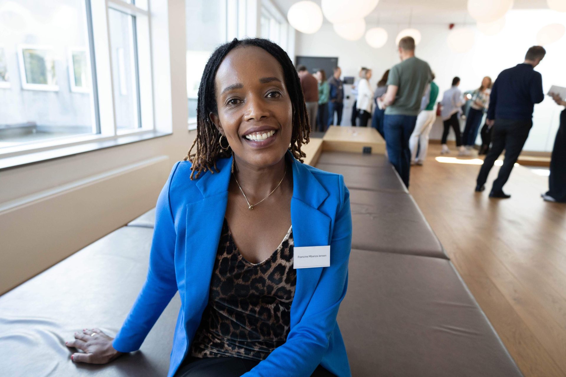 Francine Mbanza Jensen, a smiling woman in blue blazer, sits on a bench in a bright room.