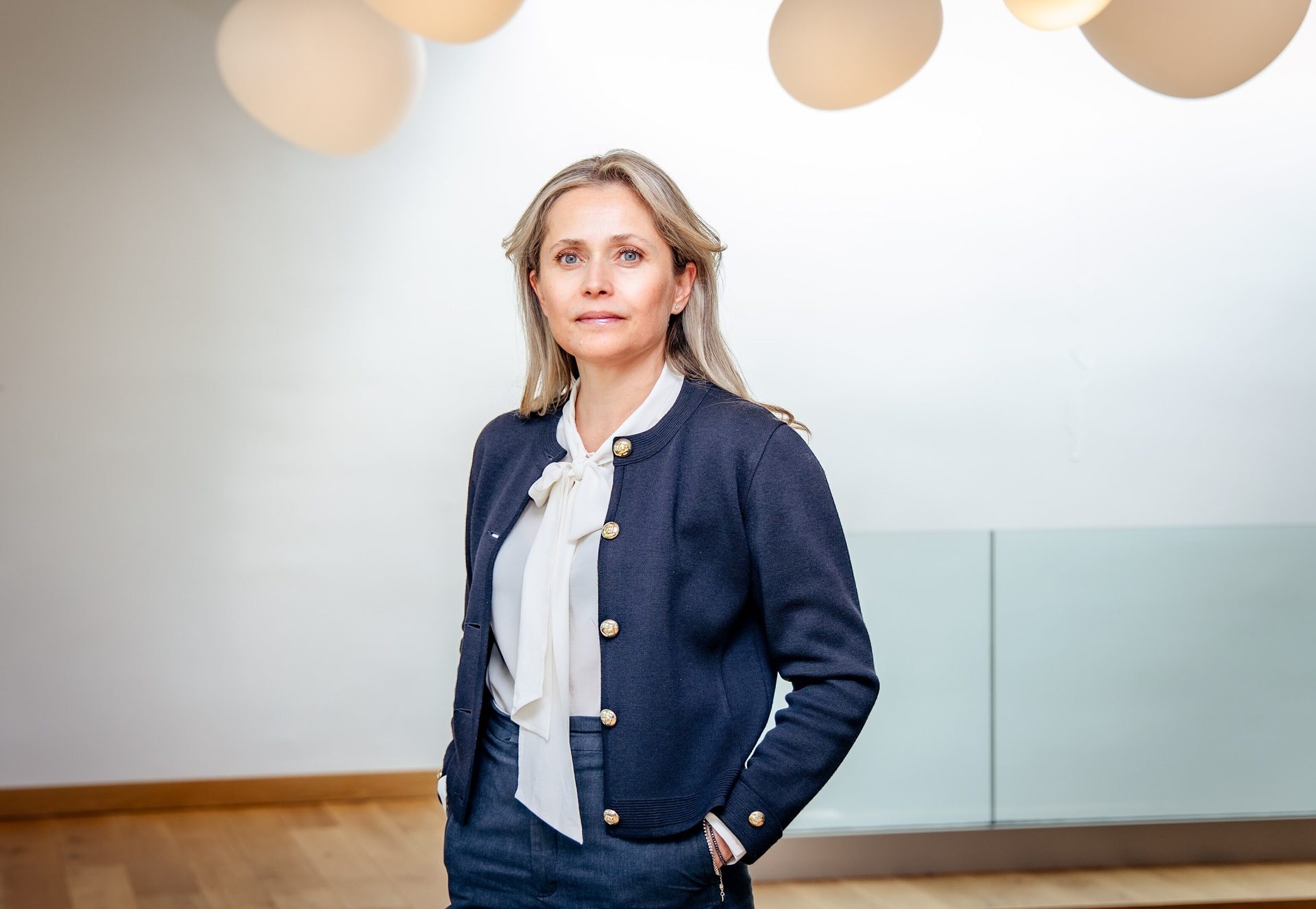 Blonde woman in white blouse and navy cardigan standing in a modern office.