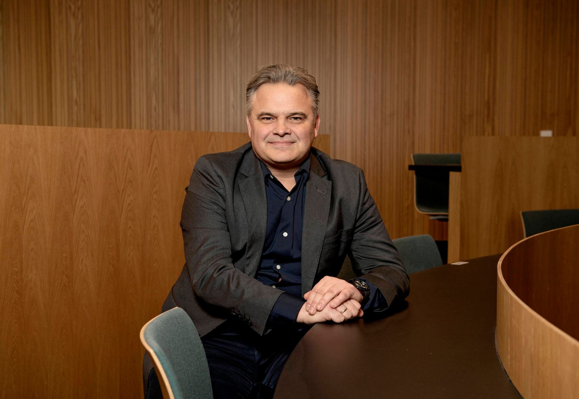 A smiling man in a dark suit jacket sits at a table in a wood-paneled room.