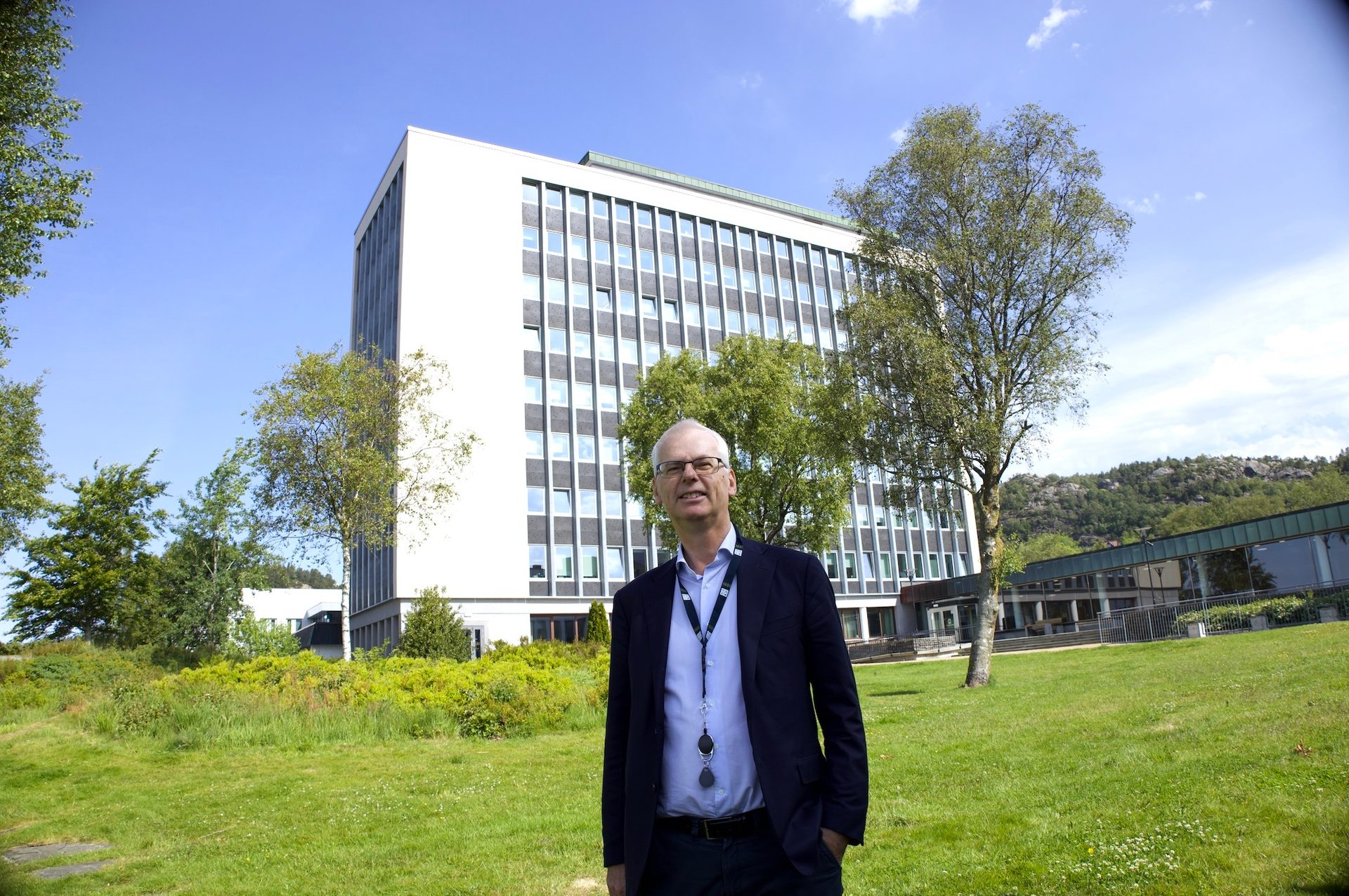 Man on green grass before a large white building, with trees and distant hills.