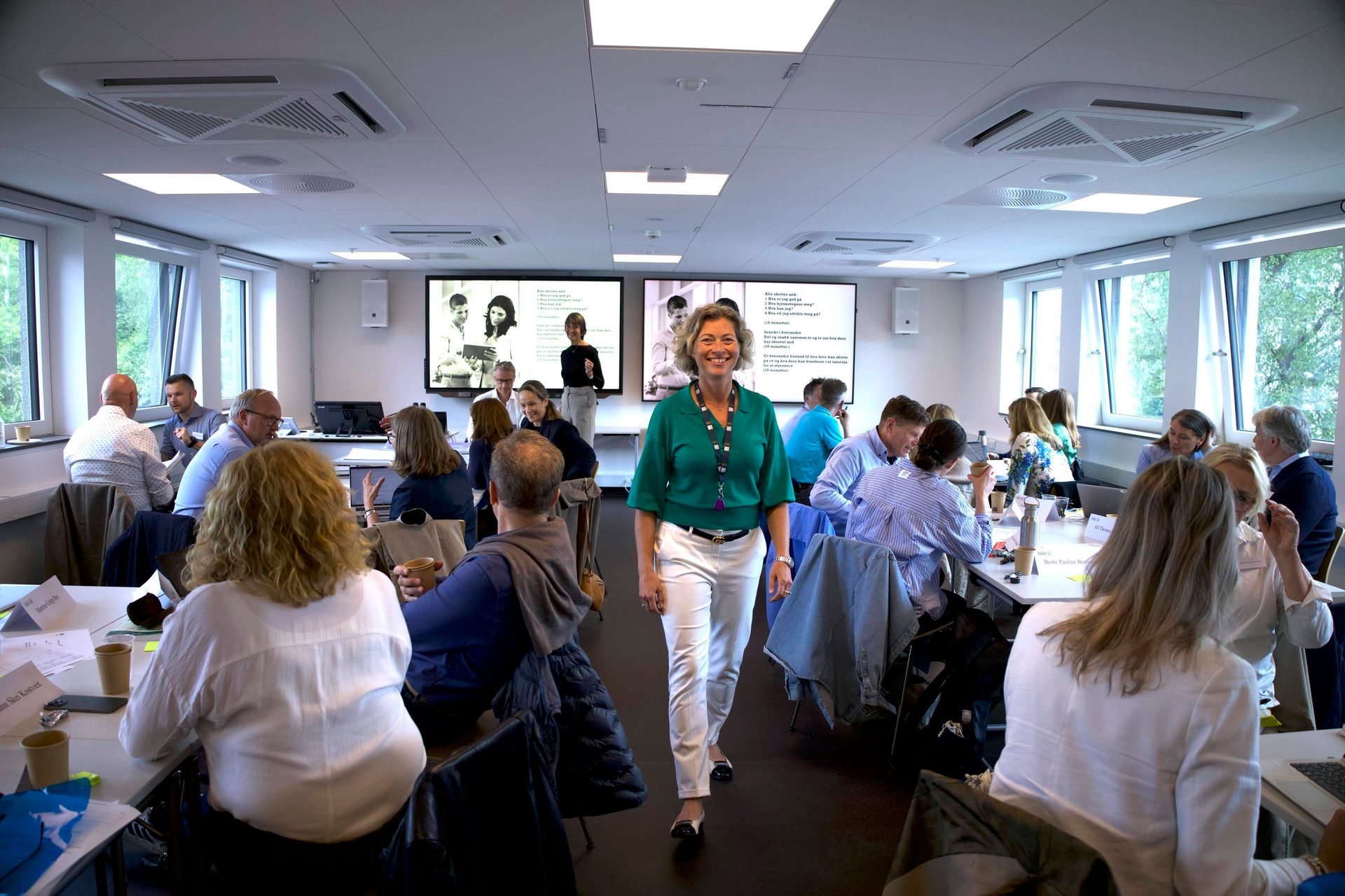 Smiling woman walks through conference room filled with attendees and screens.