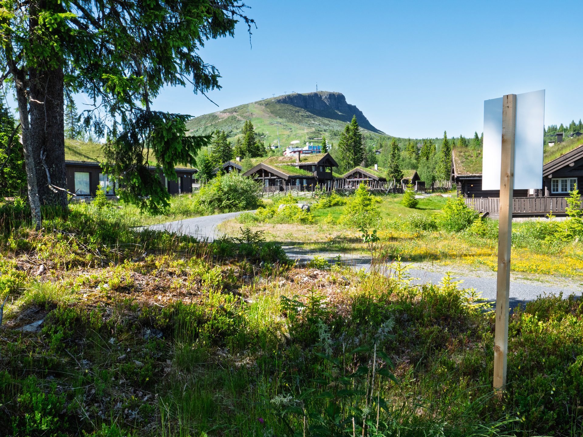 Traditional grass-roofed houses and green trees in a sunny mountain landscape, with a blank sign on the right.