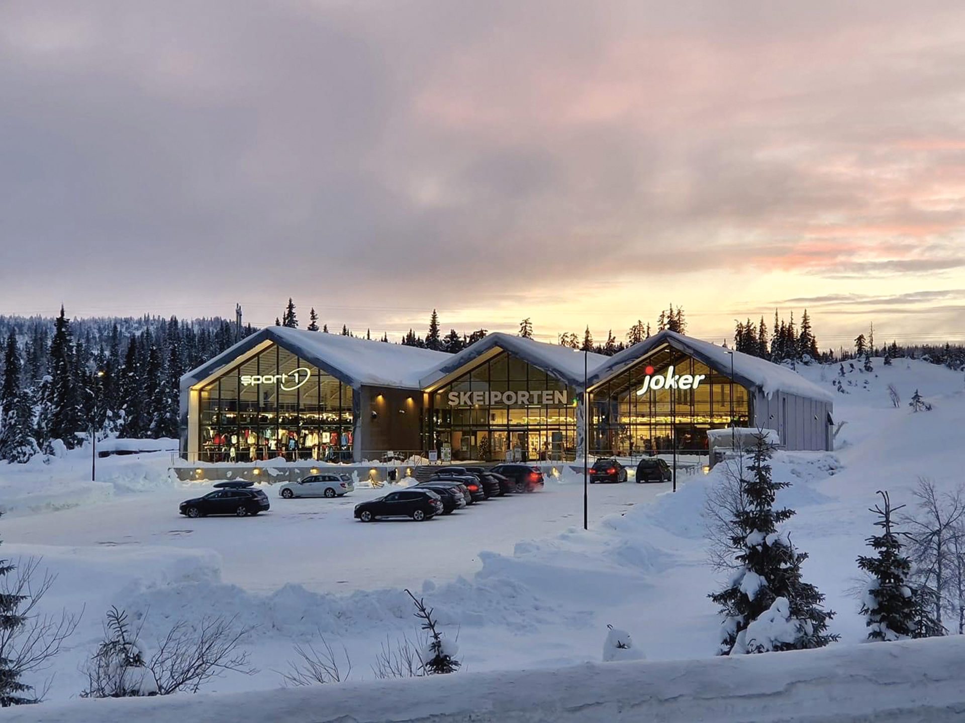Snowy shopping center (Sport 1, Skeiposten, Joker) in a winter forest, cars parked at sunset.