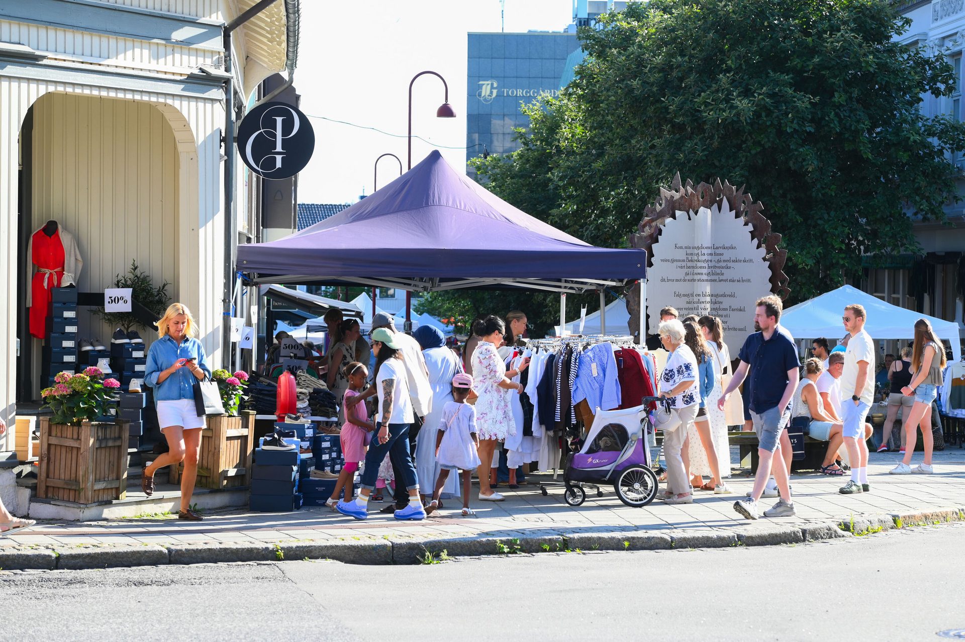An outdoor market with people browsing clothes under a purple tent on a sunny day, storefronts visible.