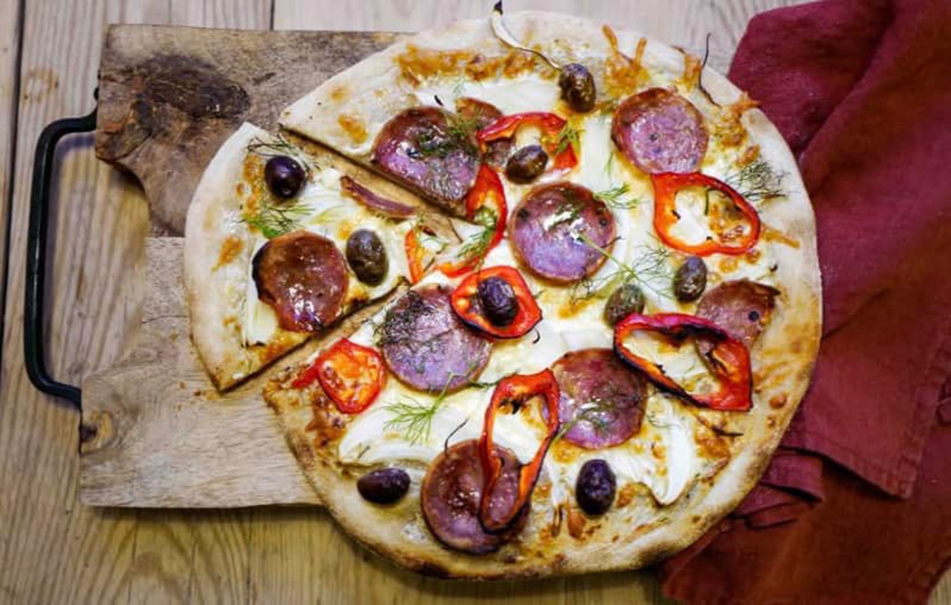 Pizza with sausage, red peppers, olives, and fennel, a slice removed, on a wooden board.